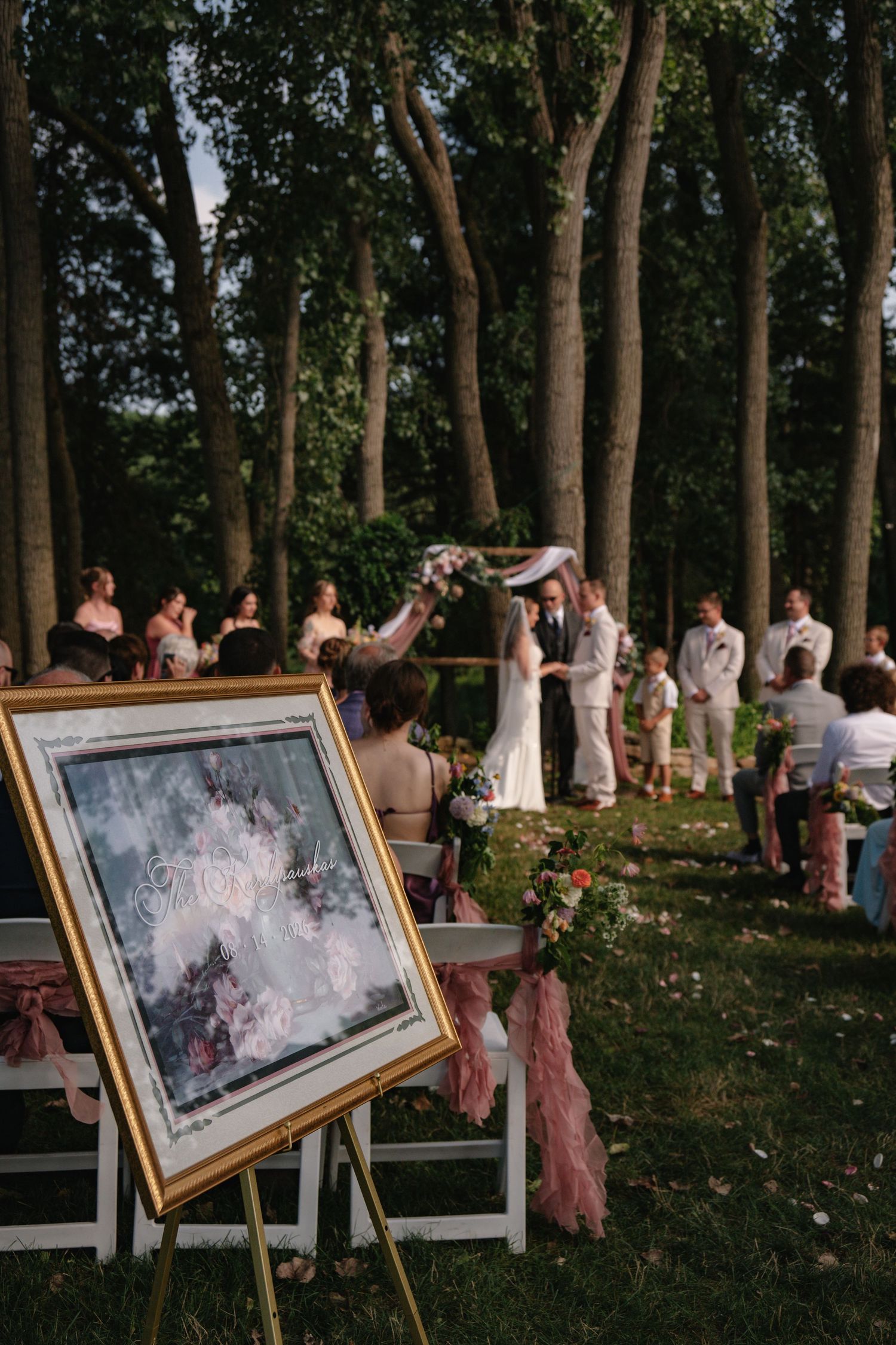 Outdoor wedding ceremony in a wooded setting with couple at floral arch, guests seated, and elegant framed welcome sign in foreground.
