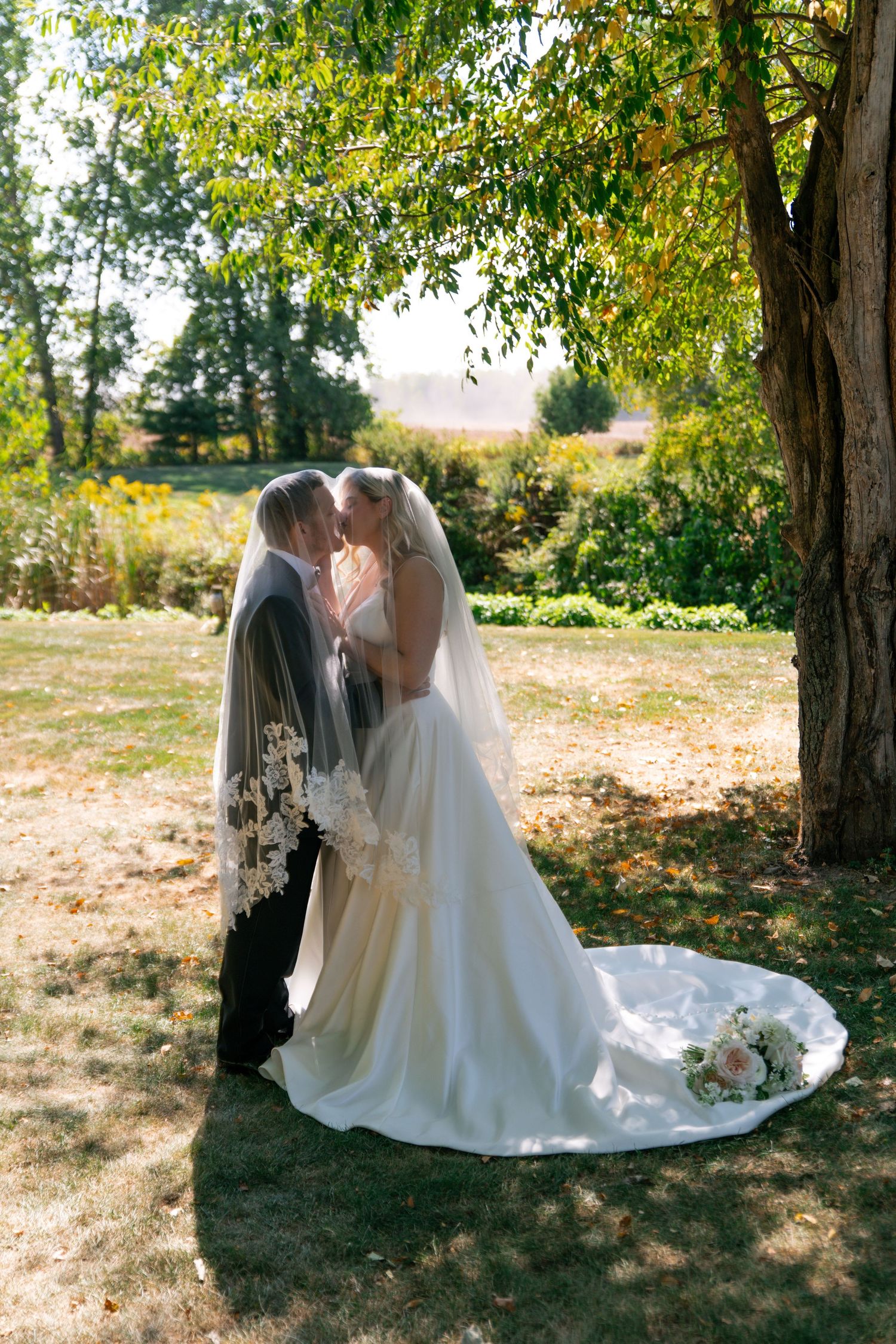 A romantic wedding couple shares an intimate moment under autumn trees in a rustic outdoor setting.
