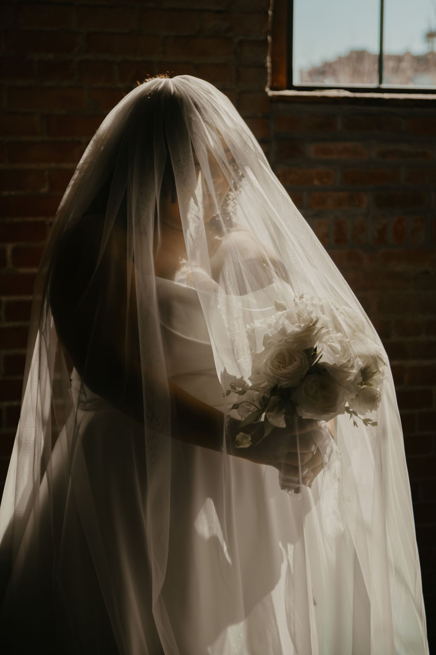 Bride in white gown and flowing veil holding white rose bouquet, standing by brick wall window in soft light.