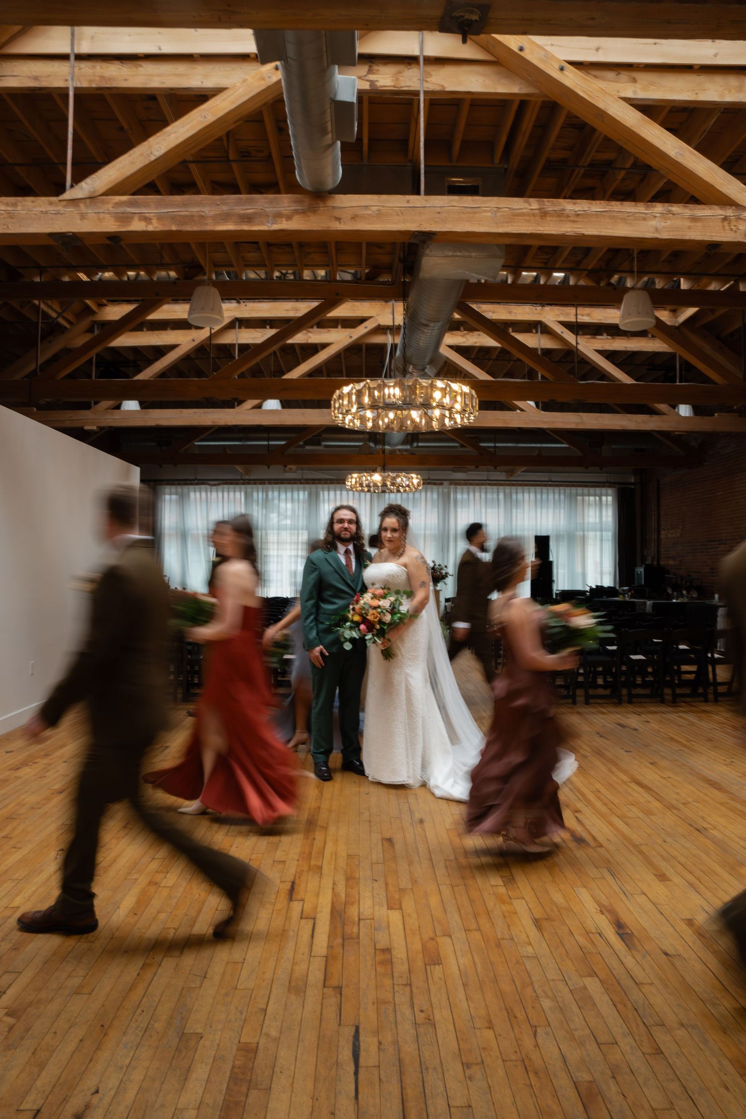 Bride and groom pose in rustic wood-beamed venue while wedding party moves around them on hardwood floor.