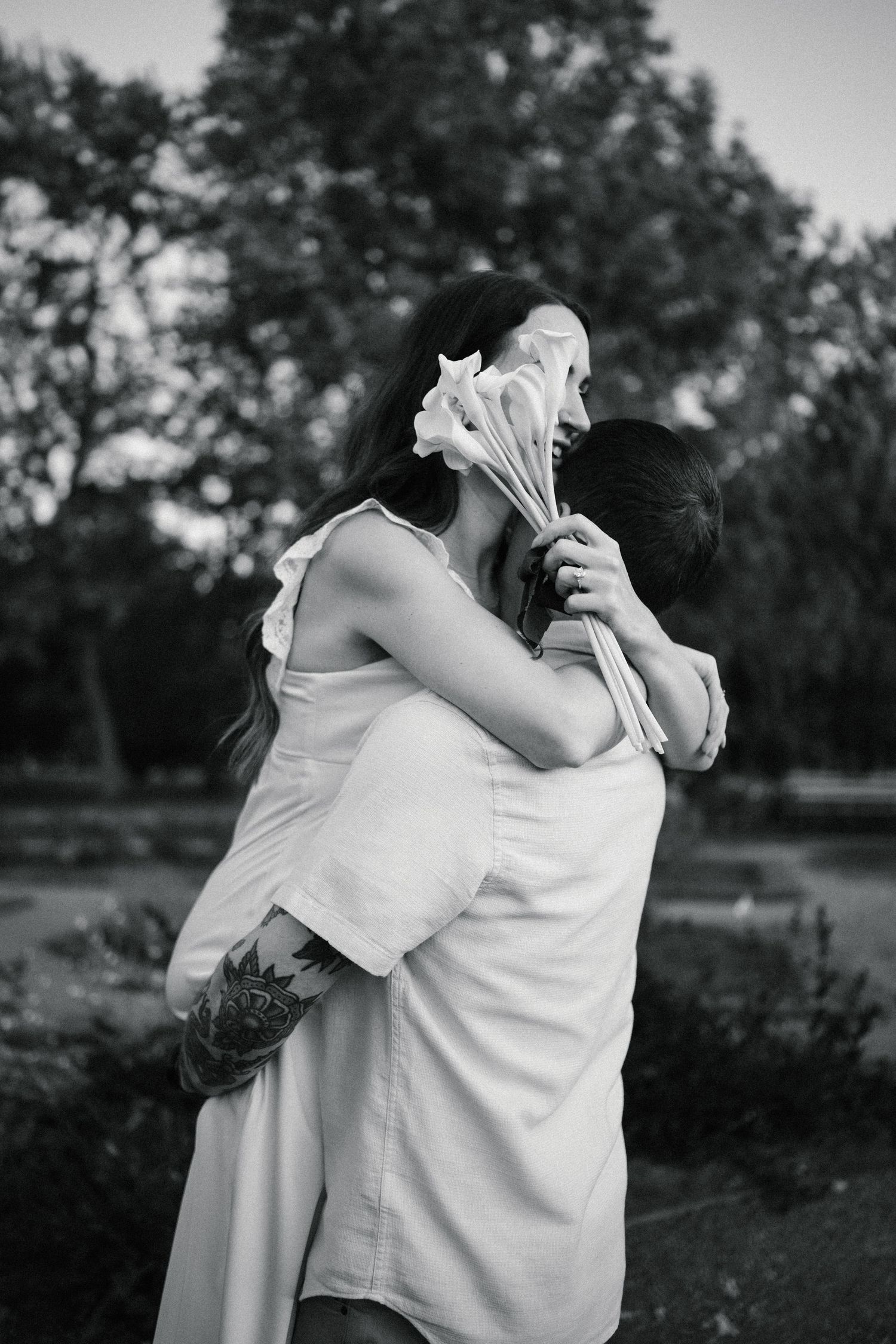 Black and white photo of couple embracing in park, woman in white dress holding calla lilies, man with tattooed arm hugging her.