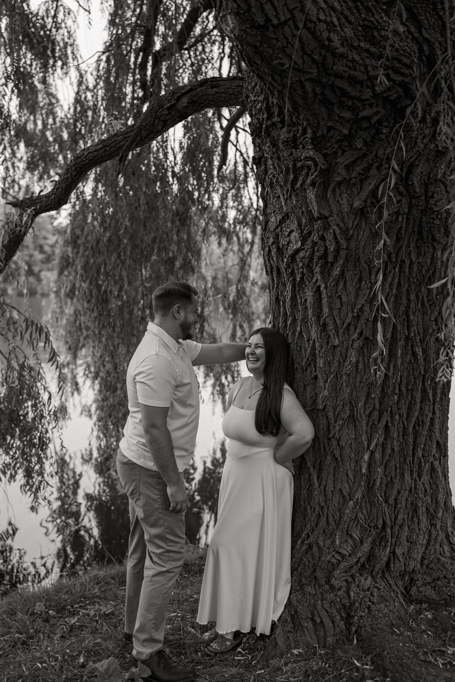 Black and white photo of a couple holding hands under a large willow tree near a lake.