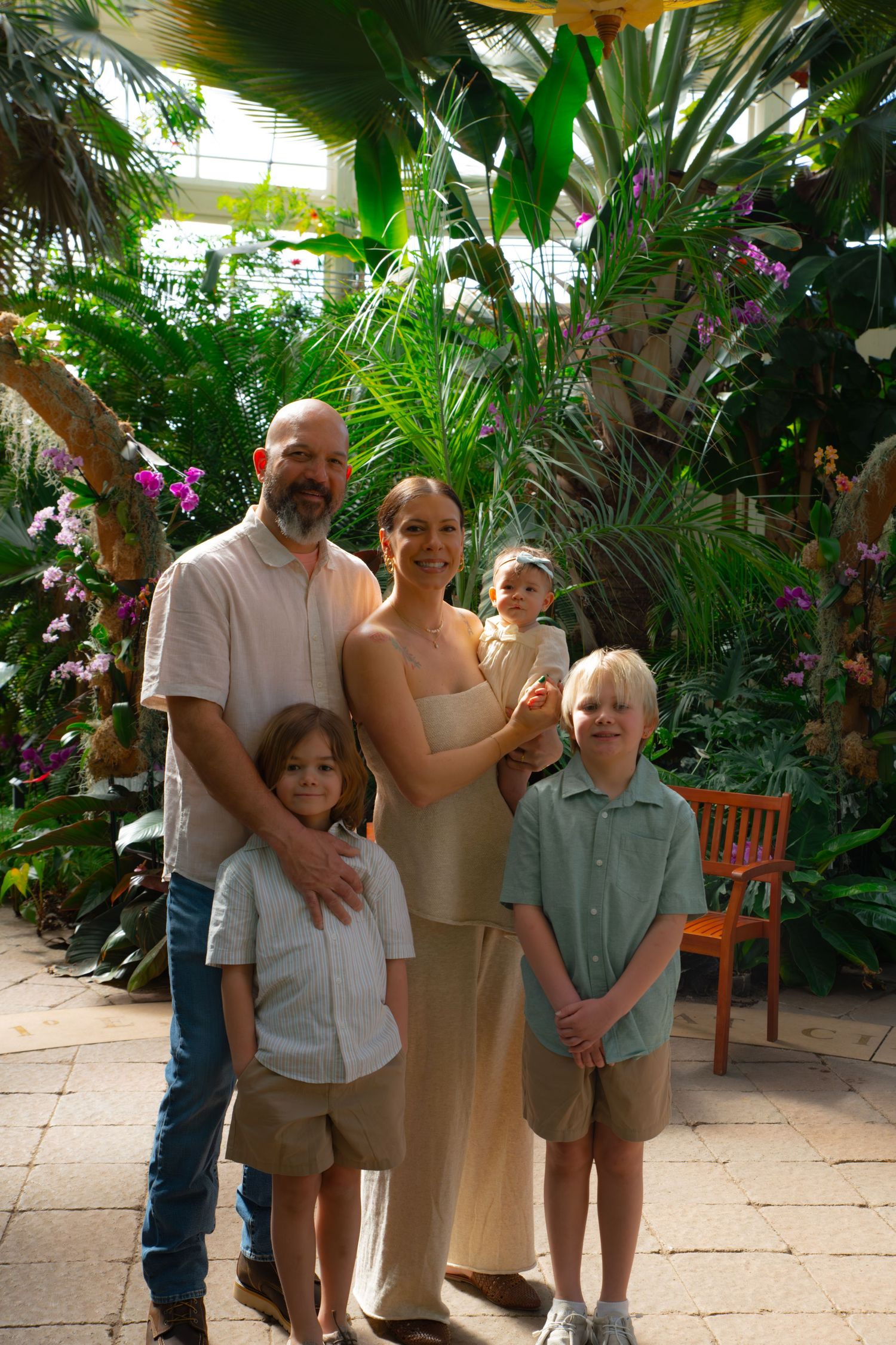 Family of five posing together in a lush tropical greenhouse with colorful orchids and green plants.