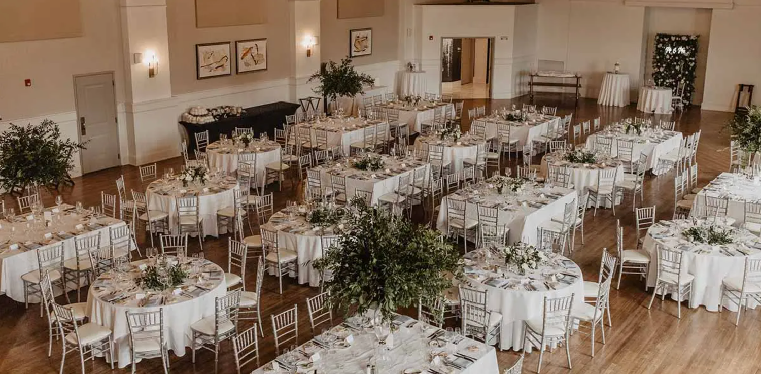 Elegant wedding reception venue with white-clothed round tables and gold chairs arranged in a spacious ballroom.