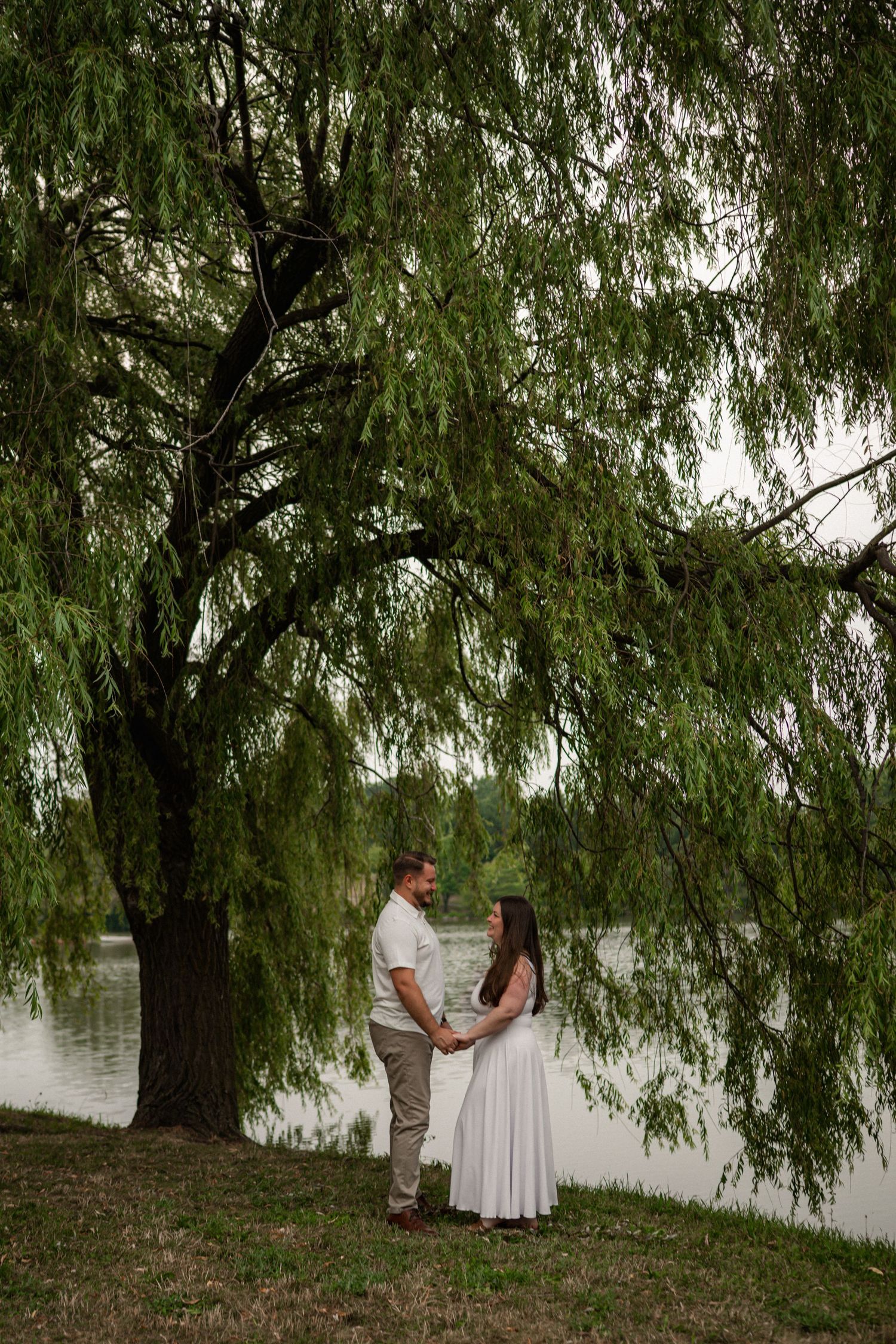 Couple holding hands under a weeping willow tree by a lake, romantic outdoor portrait.