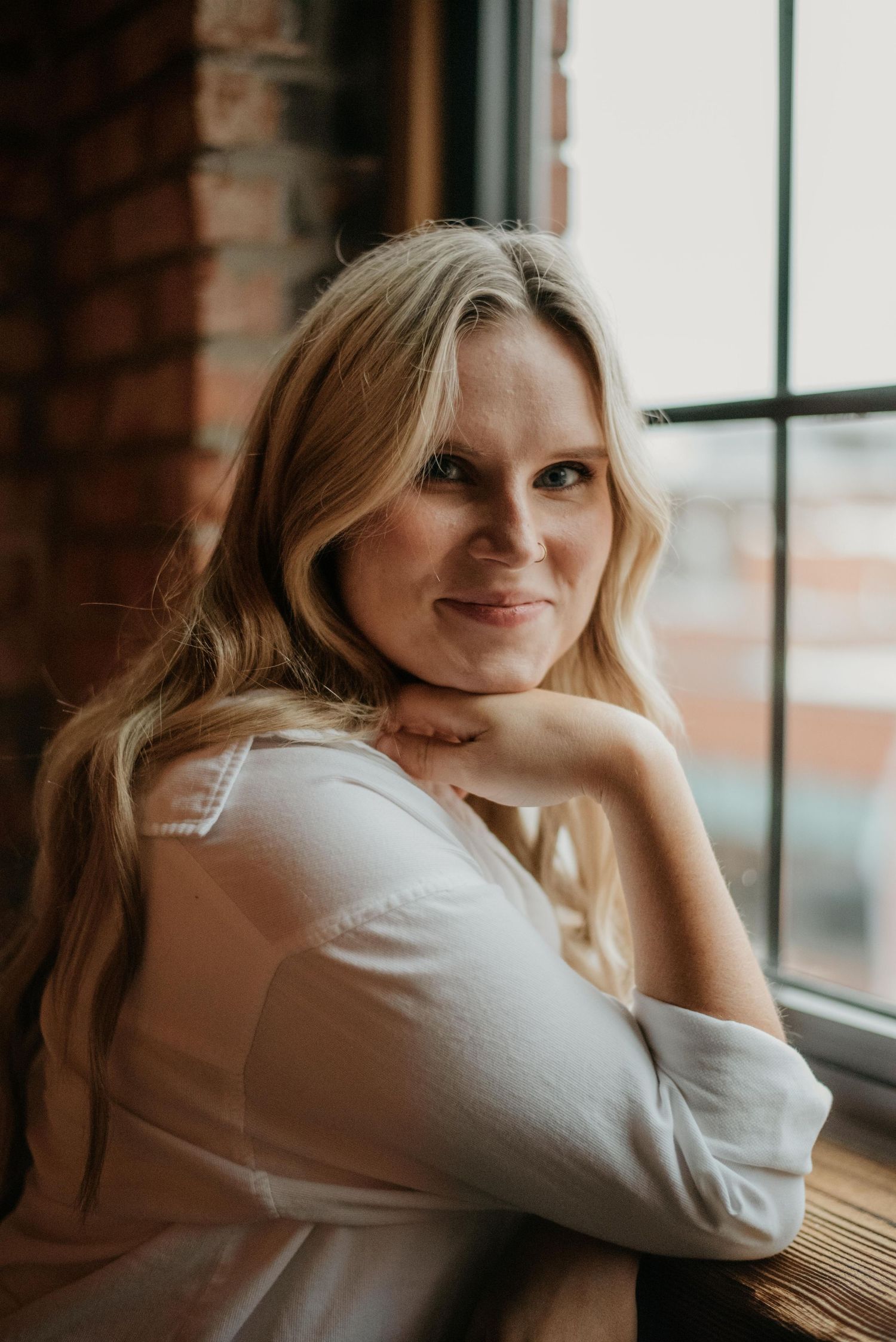 Smiling blonde woman in white top poses by brick wall window in warm indoor portrait photo.