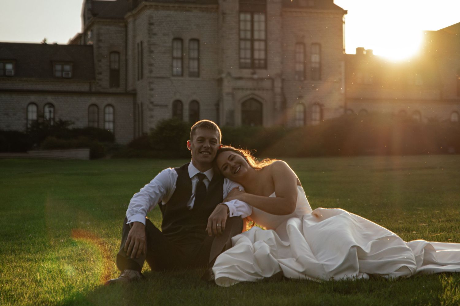 Bride and groom sitting on green grass at golden sunset hour in front of elegant historic building.