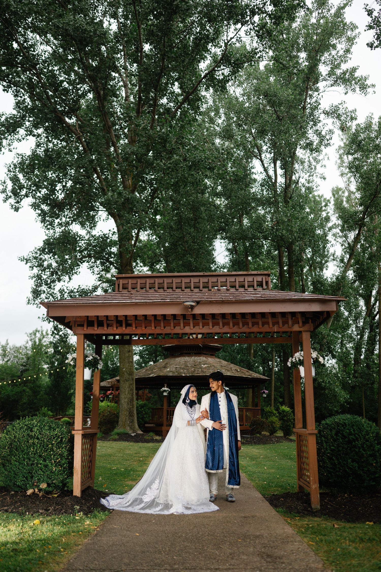 Bride and groom in traditional wedding attire standing under a wooden gazebo surrounded by lush green trees.