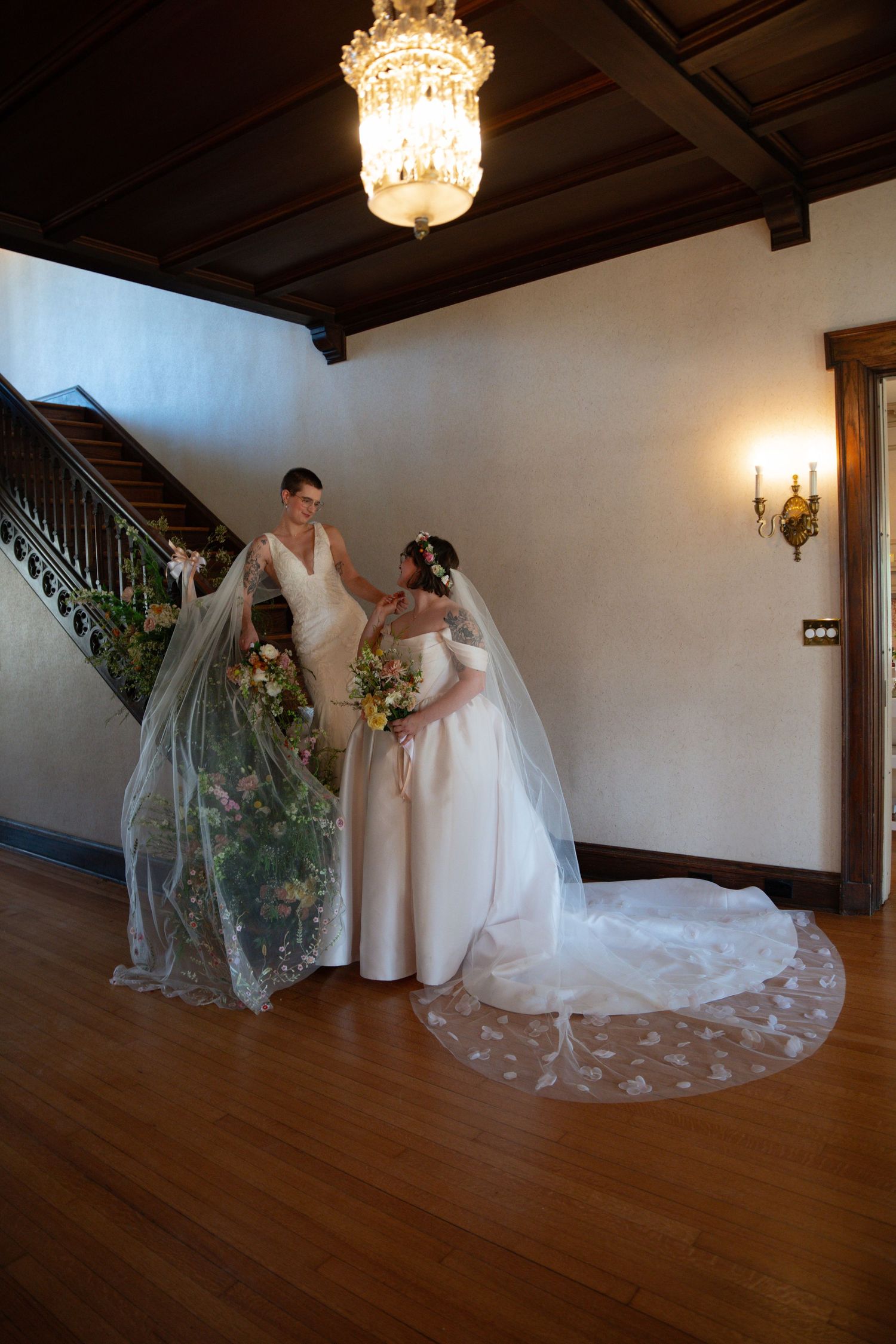 Two brides in elegant white wedding gowns with veils stand together in a grand staircase hall.