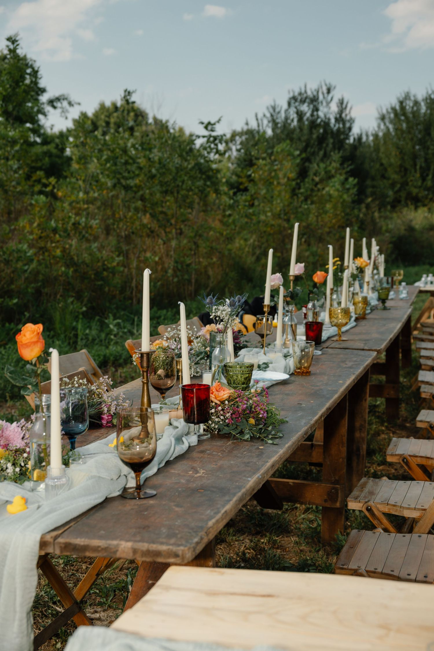 Rustic outdoor long dining table with candles, colorful flowers, and glassware set for an elegant garden wedding reception.