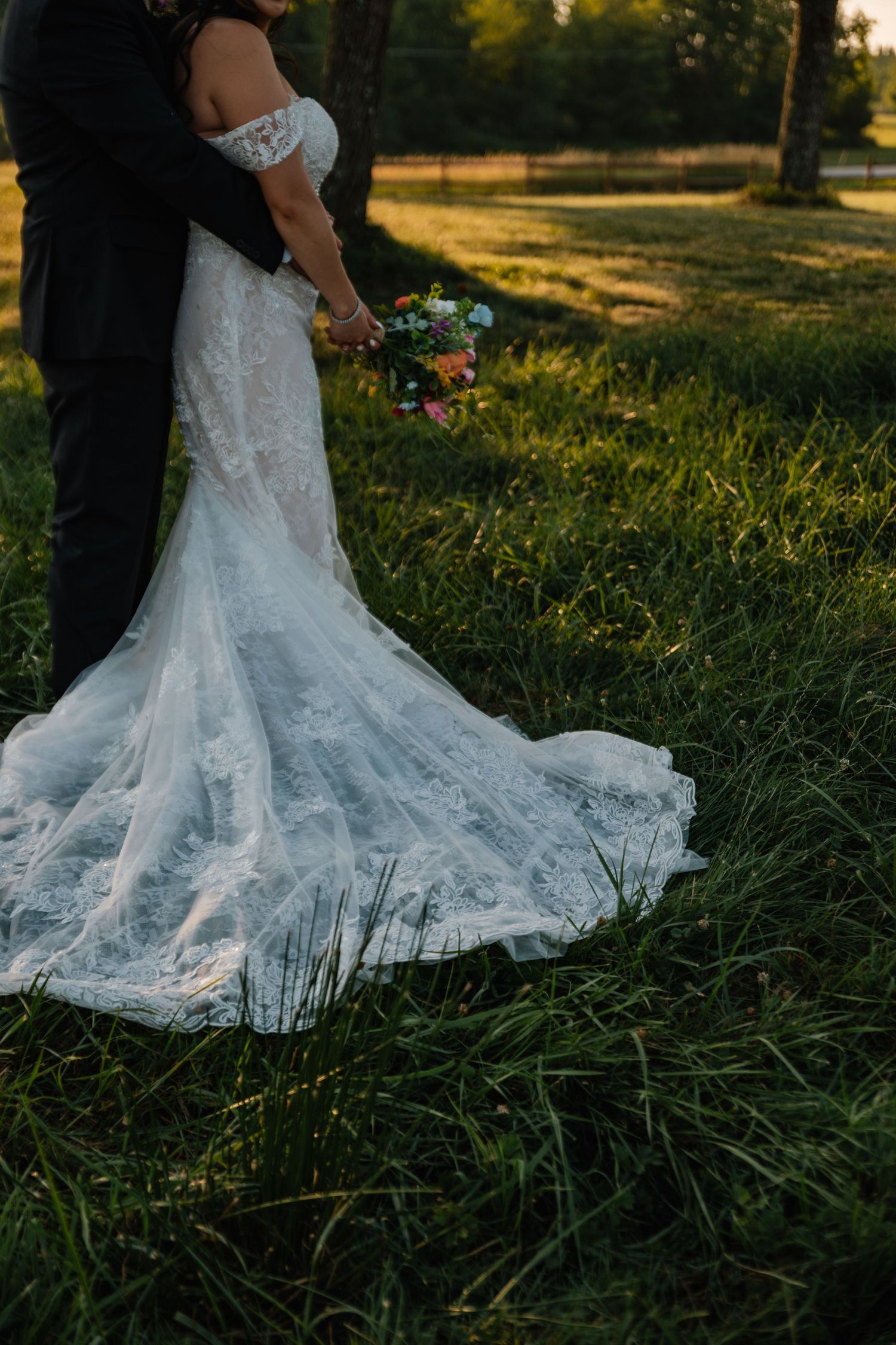 A stunning white lace wedding dress train trails on green grass during golden hour sunset in an outdoor setting.