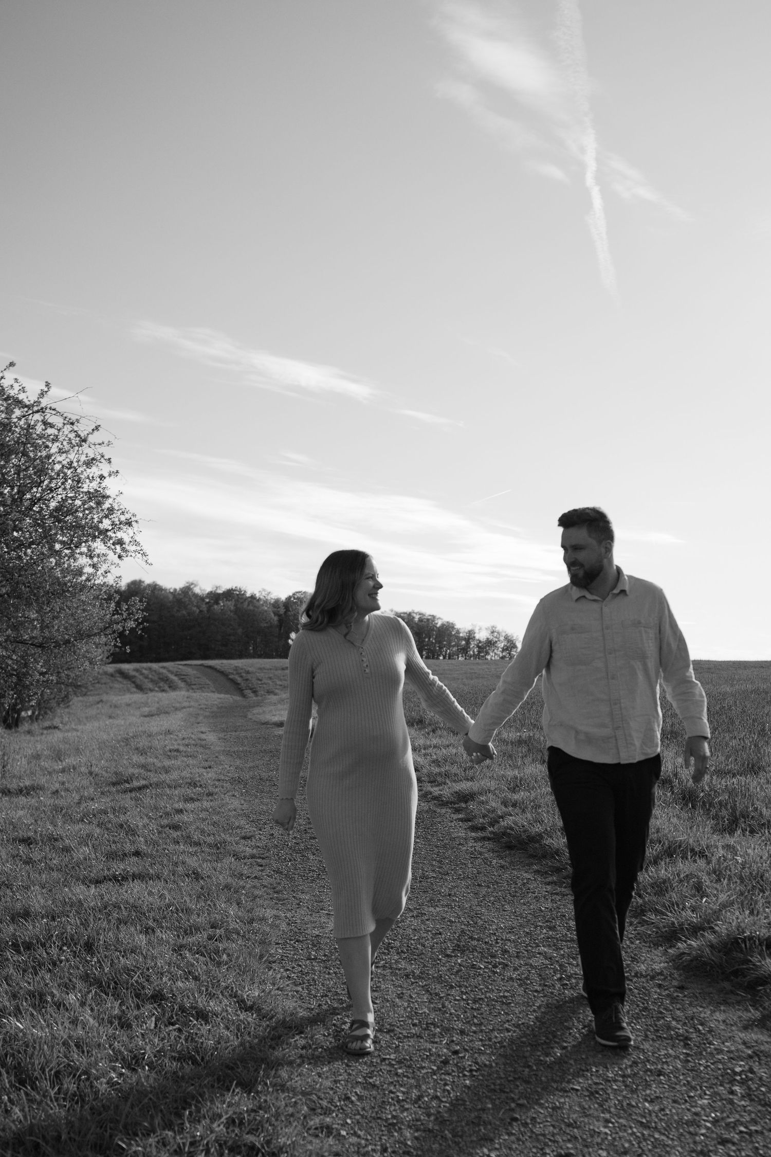 Black and white photo of a couple holding hands and walking along a rural path surrounded by fields and trees.