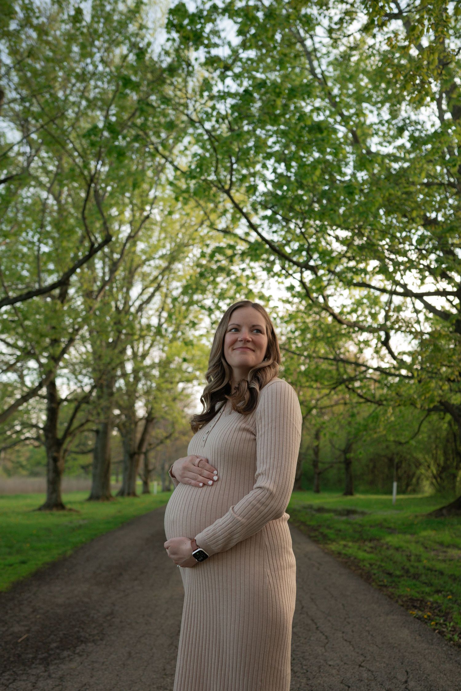 Smiling pregnant woman in beige ribbed dress cradling baby bump on tree-lined path in lush green park.