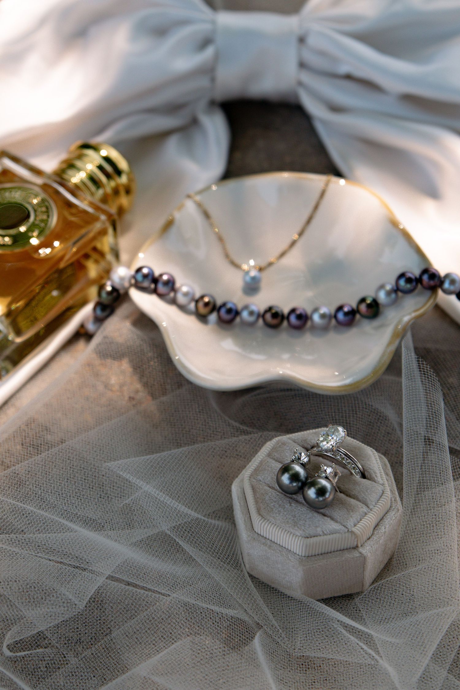 Elegant bridal jewelry flat lay with pearl necklace, diamond ring, earrings, perfume bottle, and white bow on gray surface.