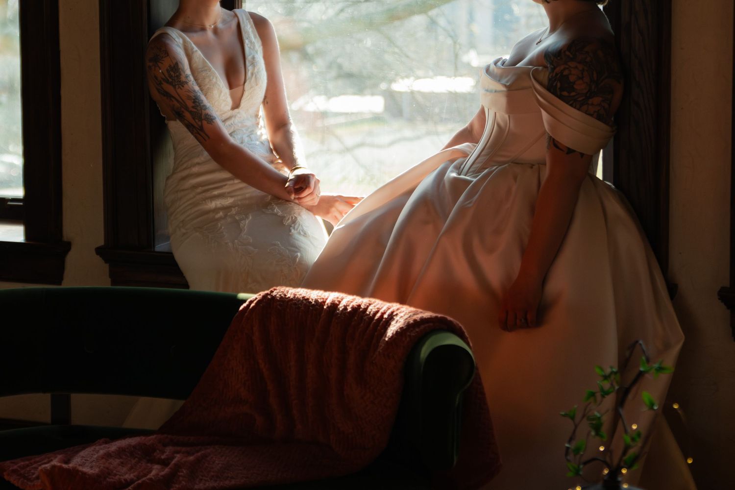 Two brides in wedding gowns share a tender moment by a sunlit window on their wedding day.