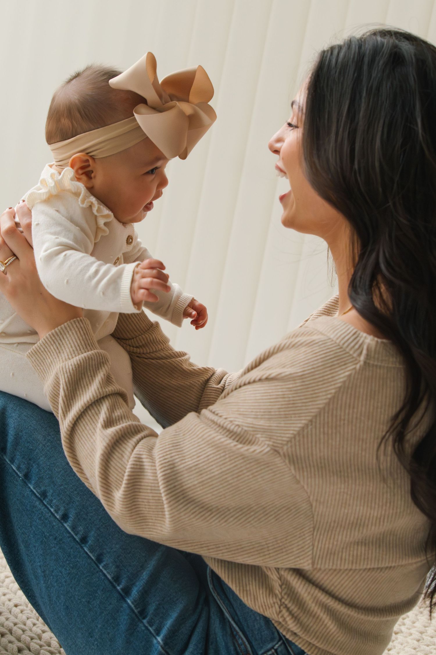 A smiling dark-haired woman in a beige sweater holds up her baby girl wearing a bow headband.