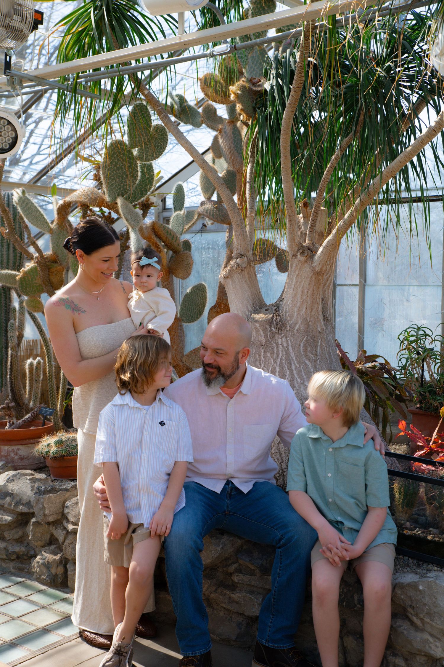 Family of five smiling together in a lush greenhouse with cacti and tropical plants.