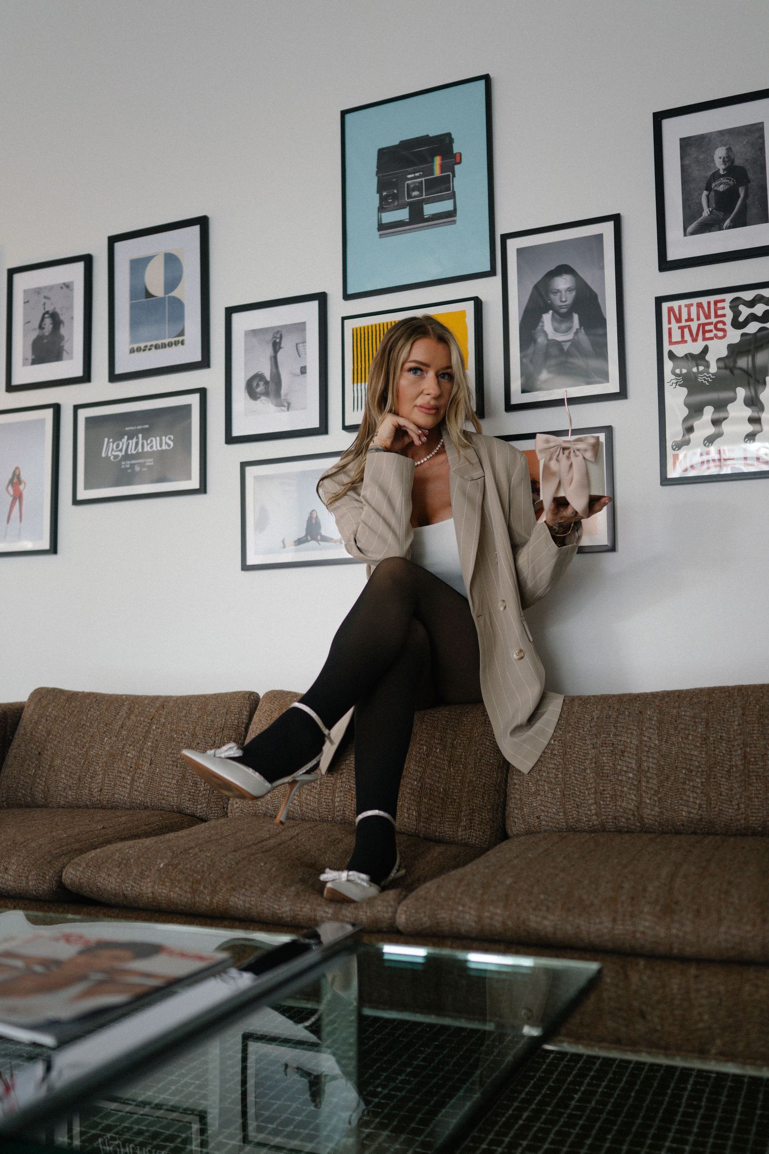 Woman in beige blazer and black leggings sitting on brown sofa with gallery wall of framed art prints behind her.