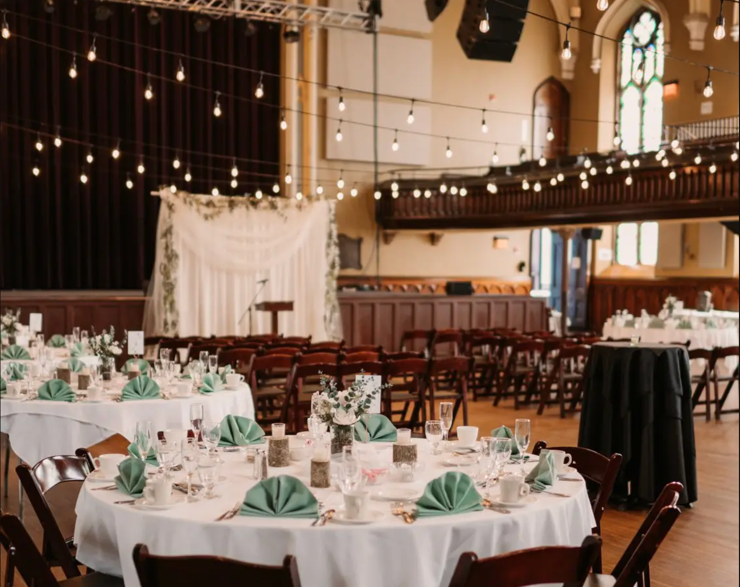 A wedding reception venue with round tables decorated with white linens and green napkins under string lights in a church hall.