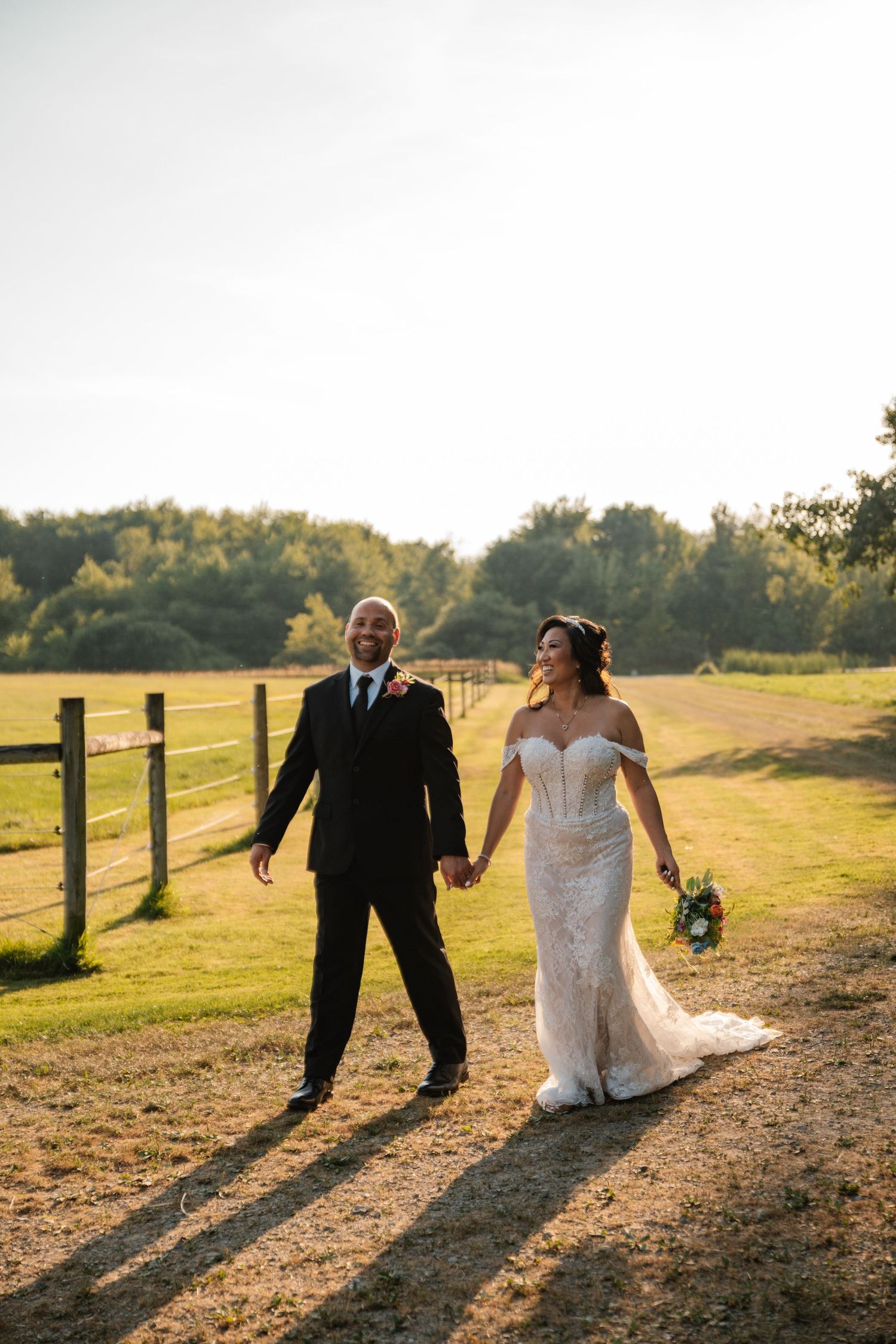 Newlywed couple walking along a sunlit path at sunset casting long shadows against a rustic fence and golden field.