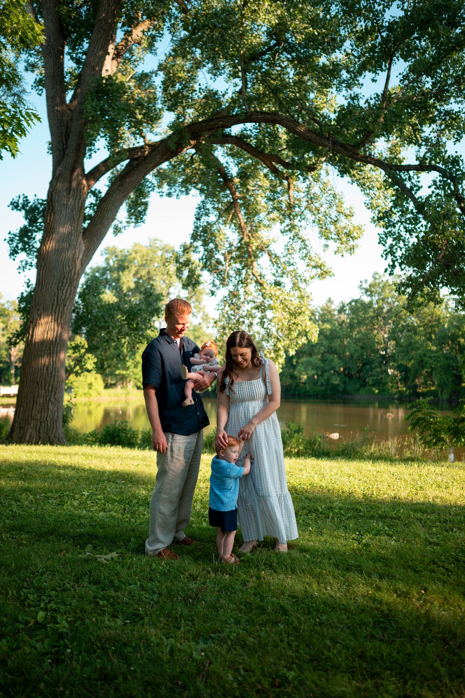 Family of four poses under large tree by pond on sunny day, mother in white dress holding toddler, father carrying baby.