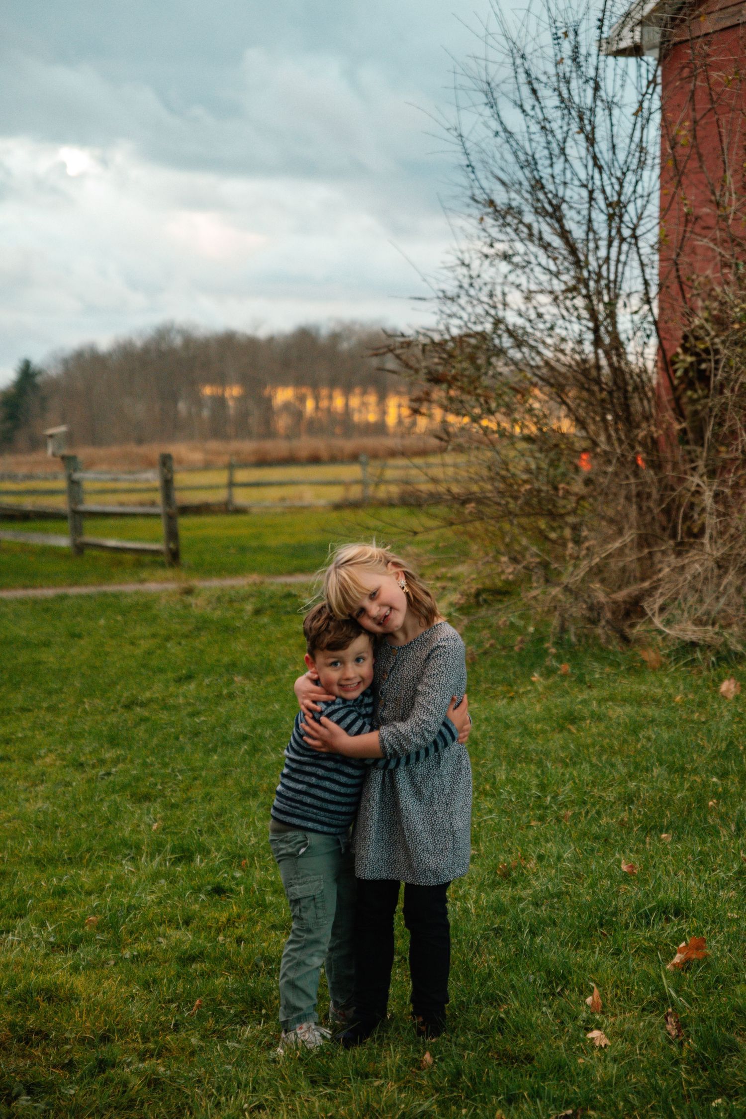 Two children hugging outdoors on a farm, smiling warmly near a red barn and wooden fence on a cloudy day.