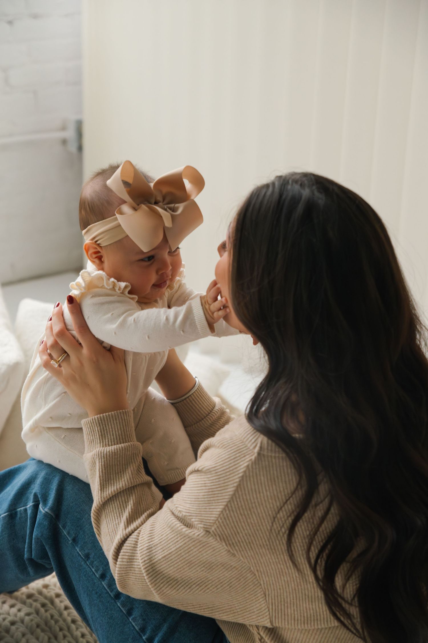A dark-haired mother holds her baby girl wearing a large bow headband, sharing a tender moment in a bright white room.