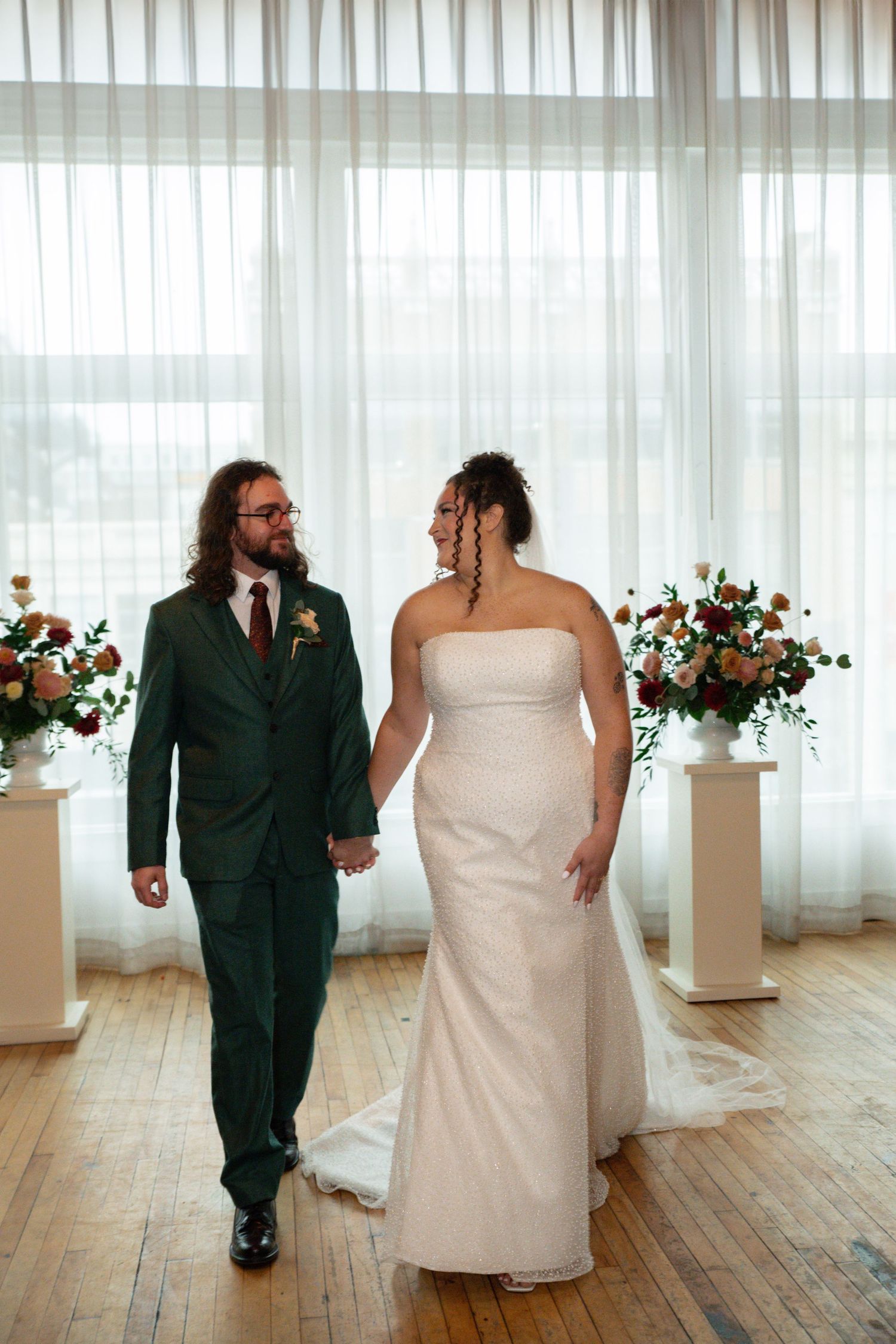 A bride in a white strapless gown and groom in a green suit hold hands walking past floral arrangements at their wedding.
