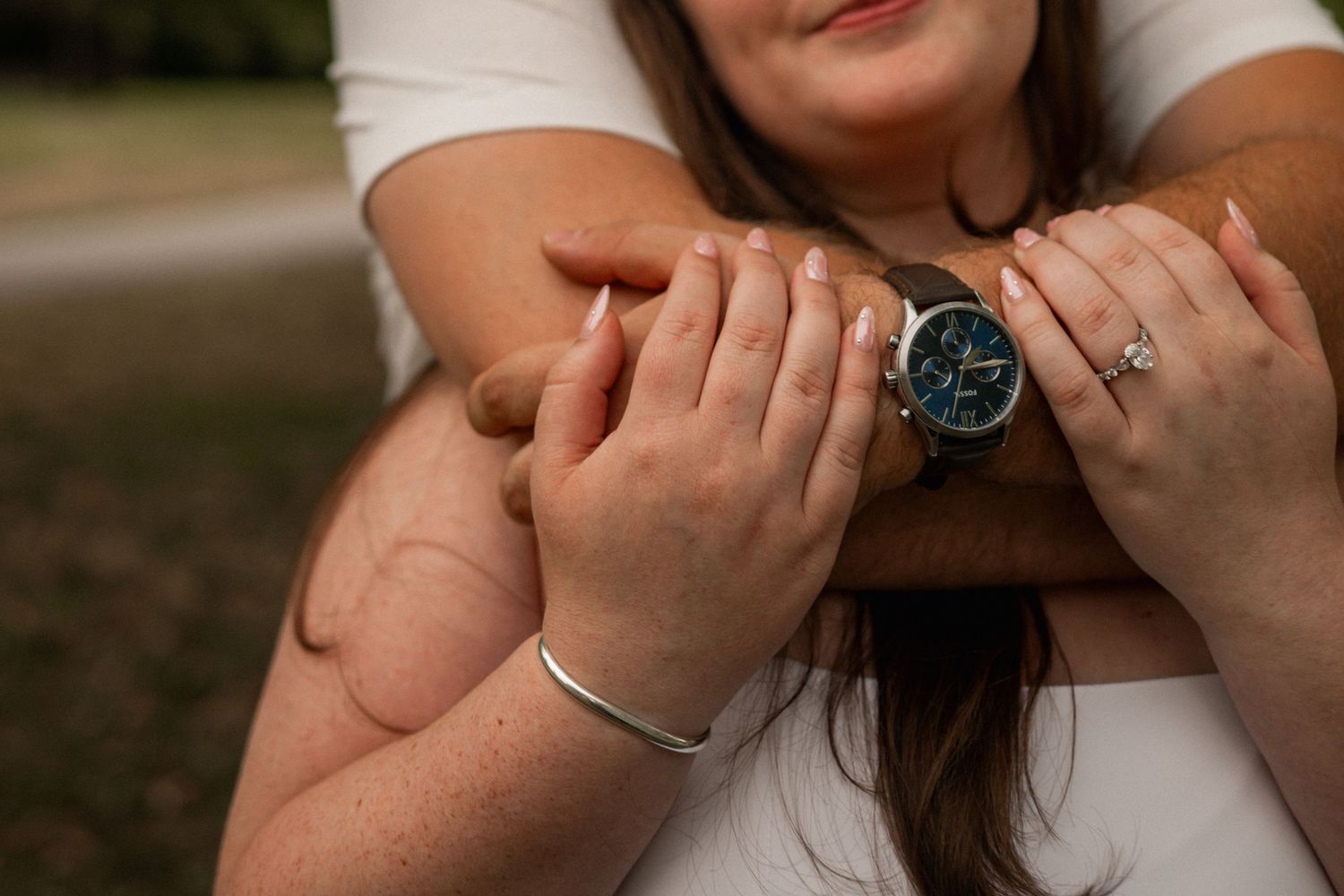 Couple holding hands showing a blue watch and diamond ring, with a silver bracelet, in an outdoor setting.