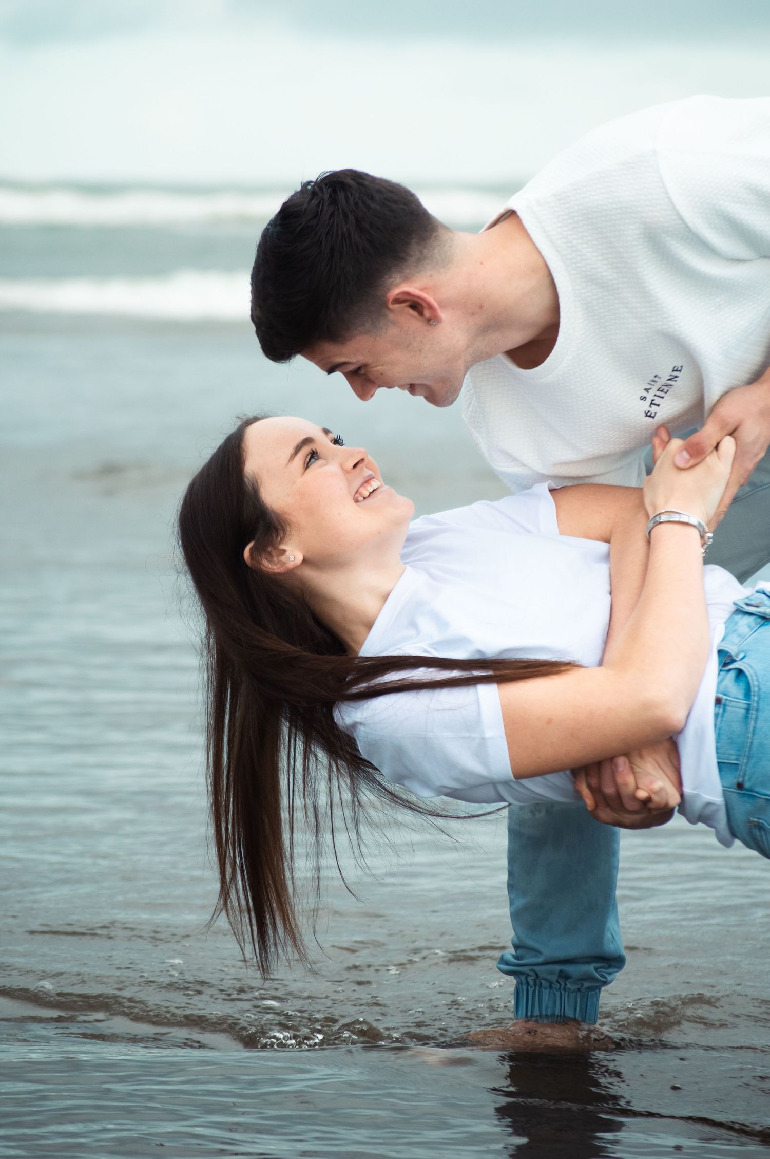 A joyful couple embraces on a beach shoreline, with the man lifting the laughing woman in his arms.