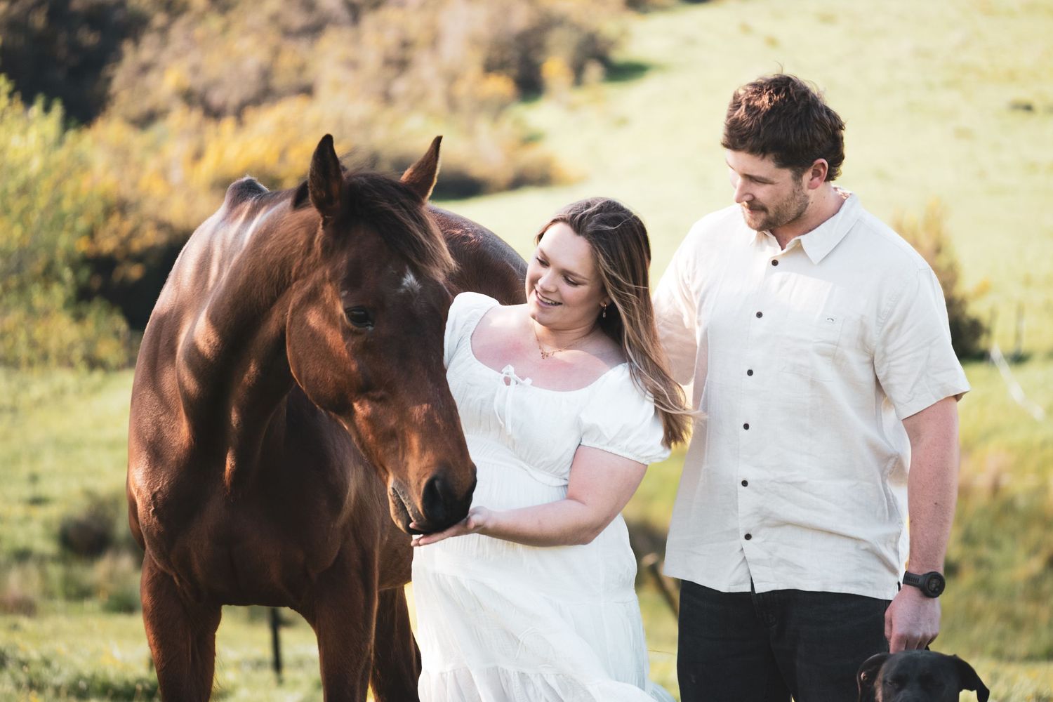 Couple in white attire stands with a brown horse in a lush green field during an outdoor engagement photo session.