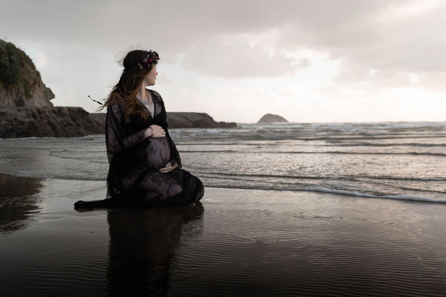 Pregnant woman in black dress sitting on dark sand beach at dusk, gazing at ocean waves with rocky cliffs behind.