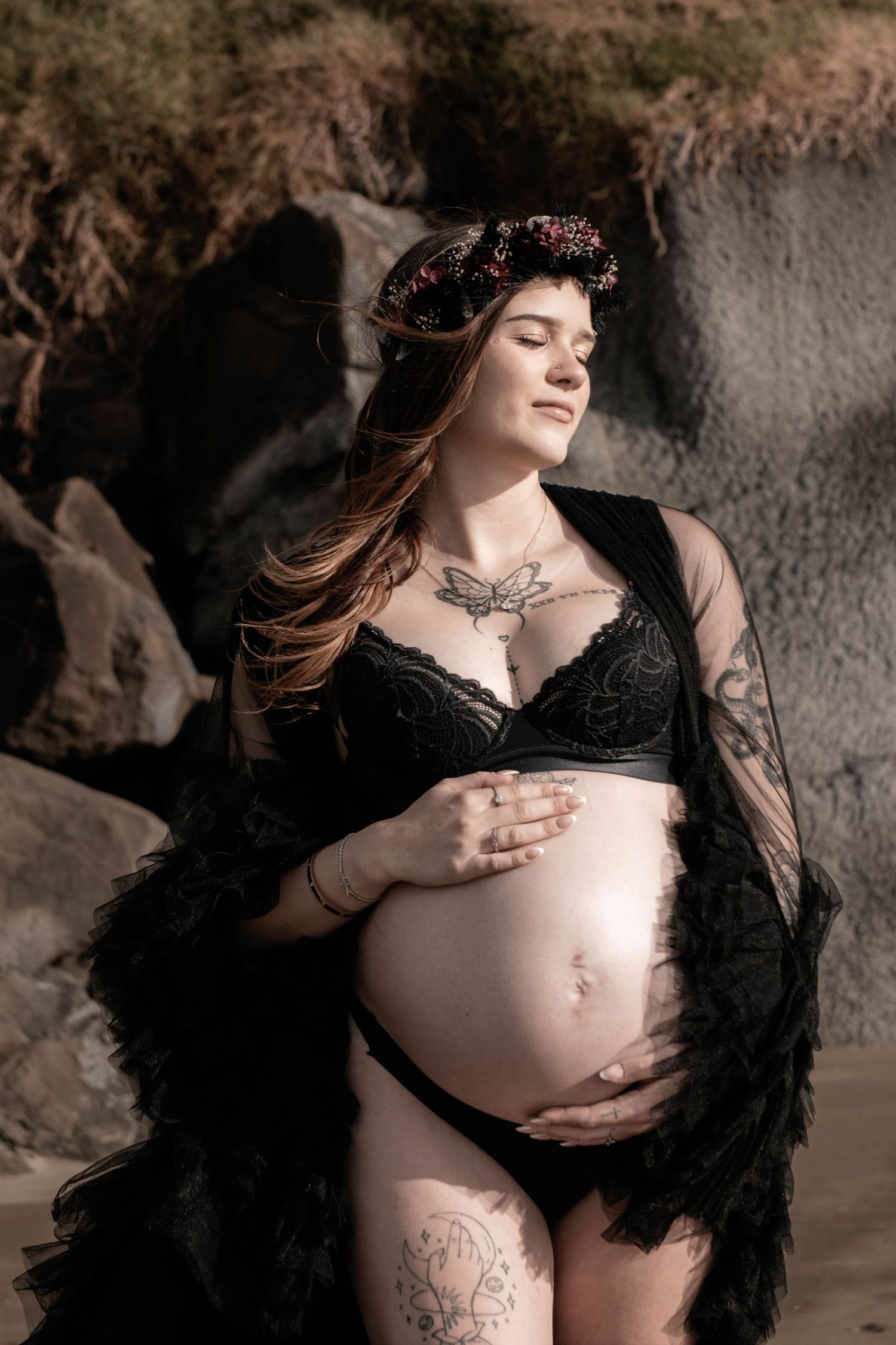 Pregnant woman in black lace lingerie and floral crown cradles baby bump outdoors near rocky backdrop.