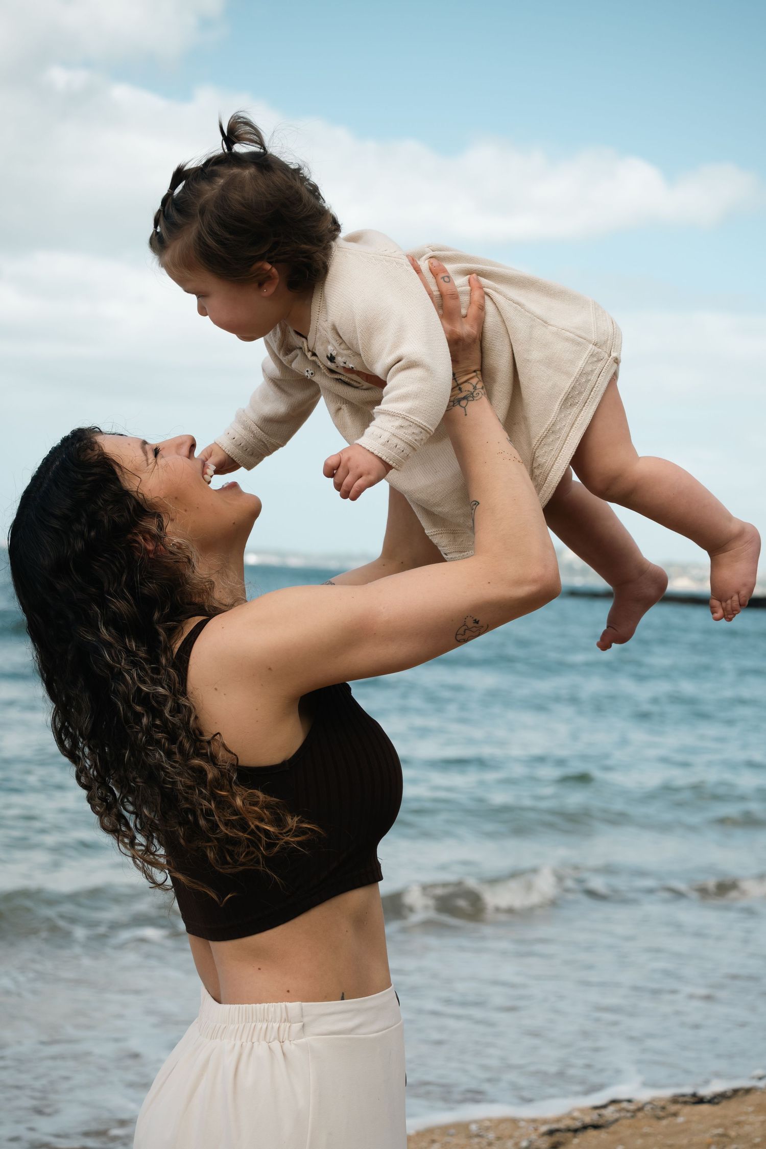 A mother joyfully lifts her toddler daughter in the air on a beach with ocean waves in the background.
