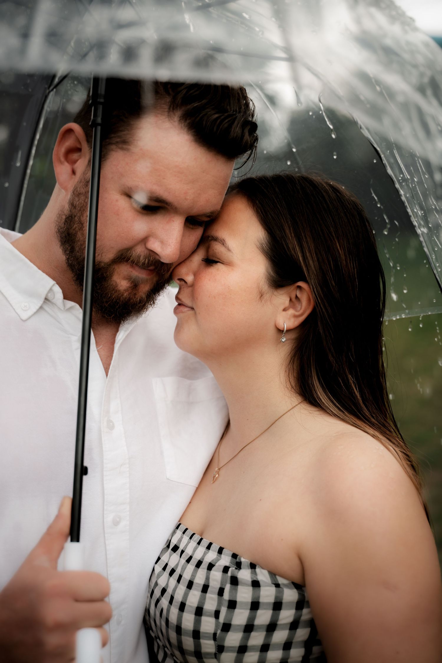 Couple sharing a tender moment under a clear umbrella in the rain, man in white shirt and woman in checkered top.