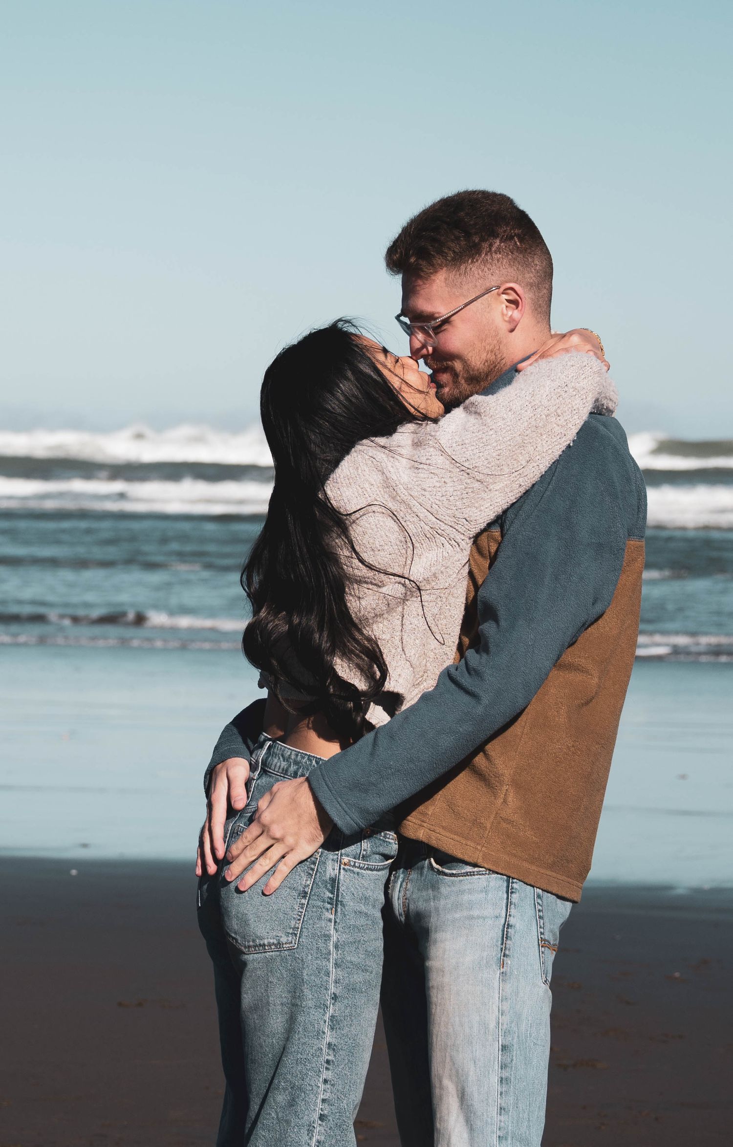 A couple embracing romantically on a sandy beach with ocean waves in the background on a sunny day.