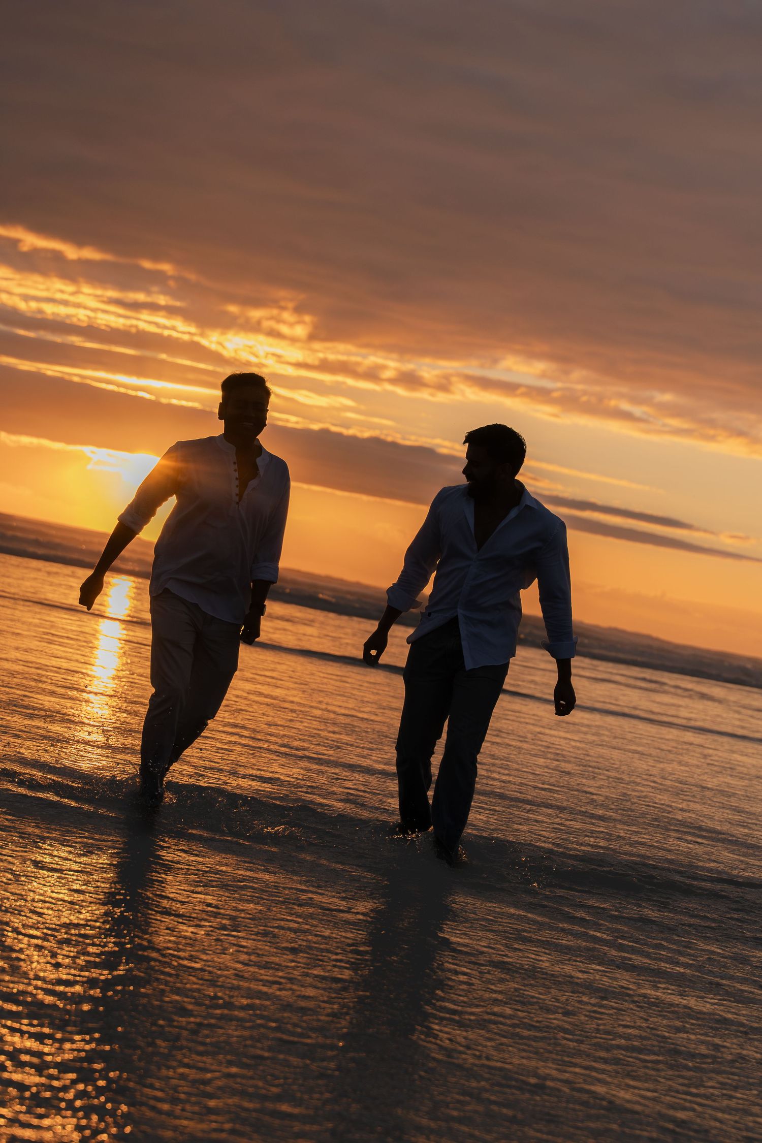 Two men walking hand in hand on a beach at sunset, silhouetted against a vibrant orange sky.