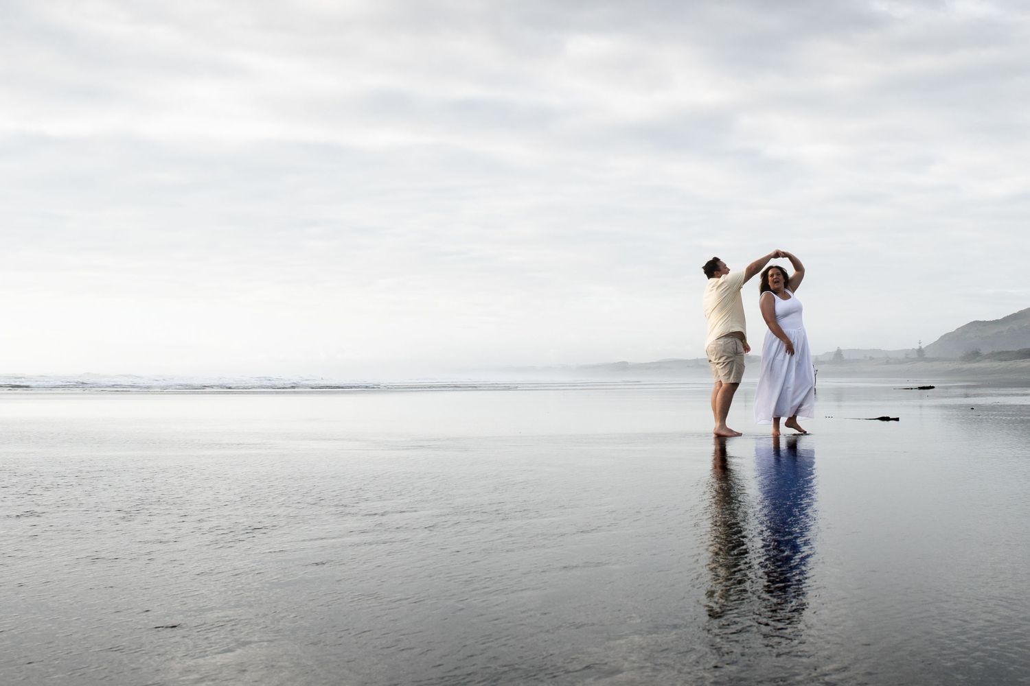 Couple dancing romantically on a calm beach shore with misty mountains and reflective water in the background.