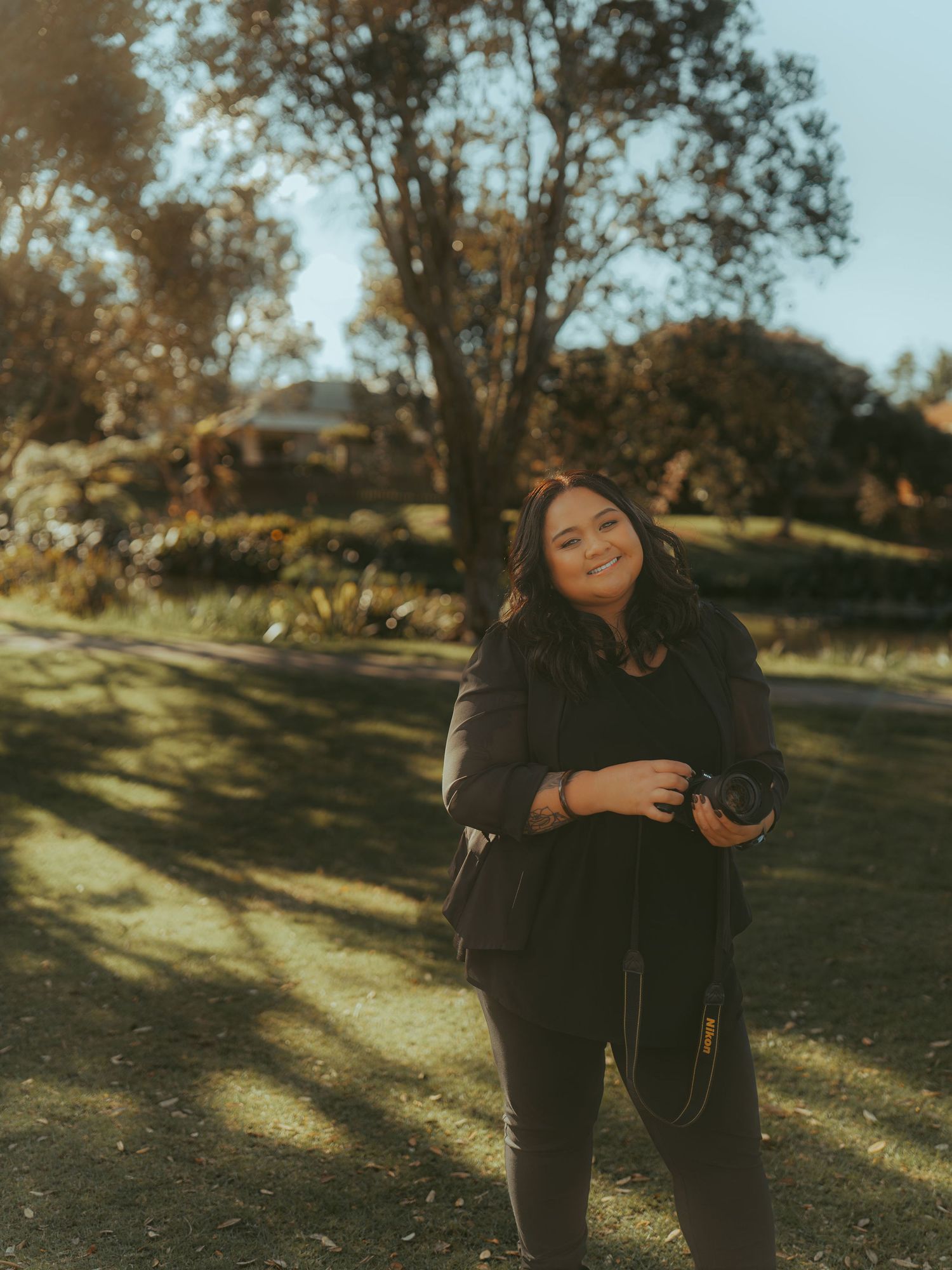 Woman in black outfit smiling outdoors in a sunlit park with trees and green grass in the background.