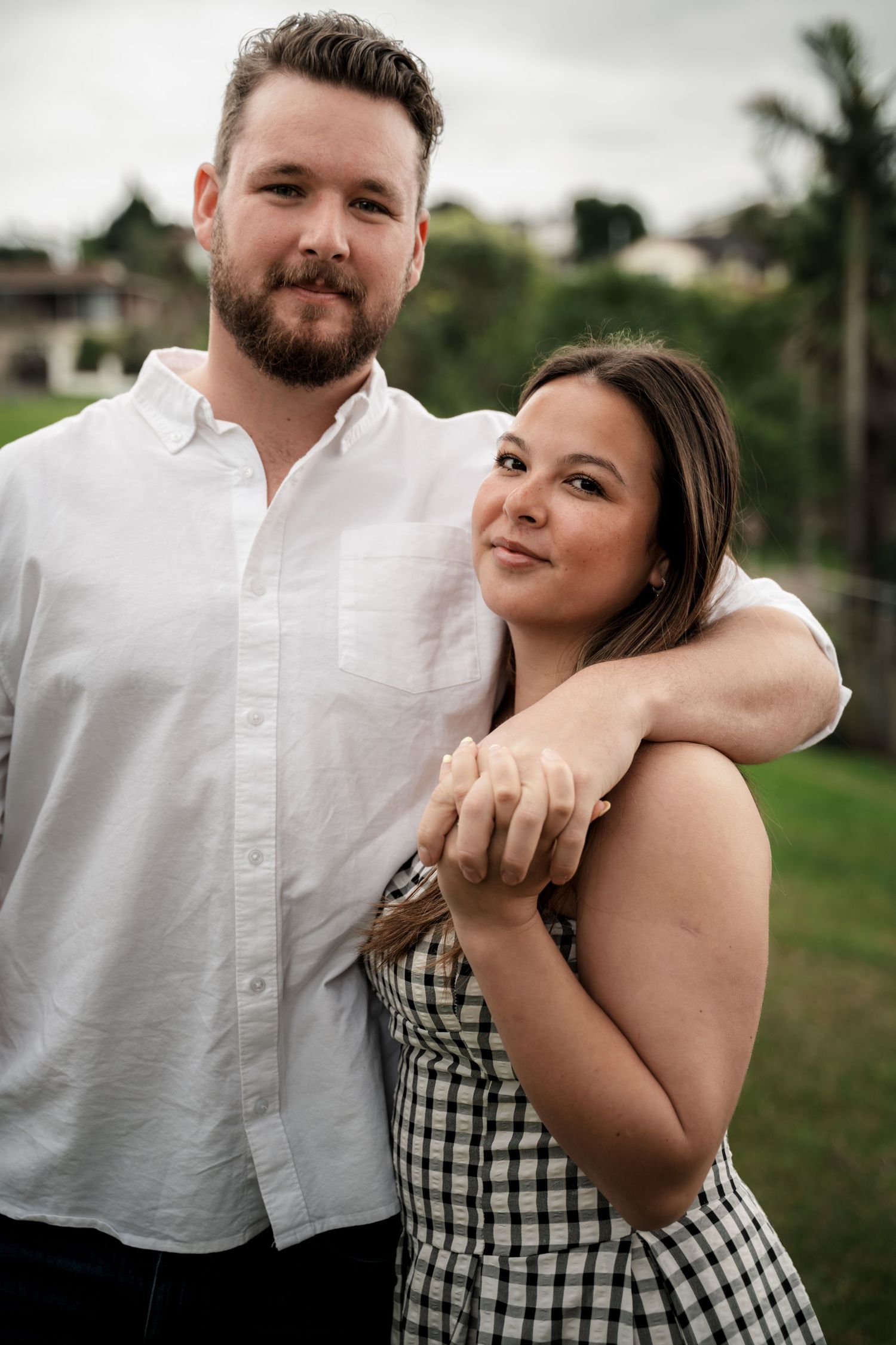 A couple poses outdoors, man in white shirt with arm around woman in checkered dress, green trees behind them.