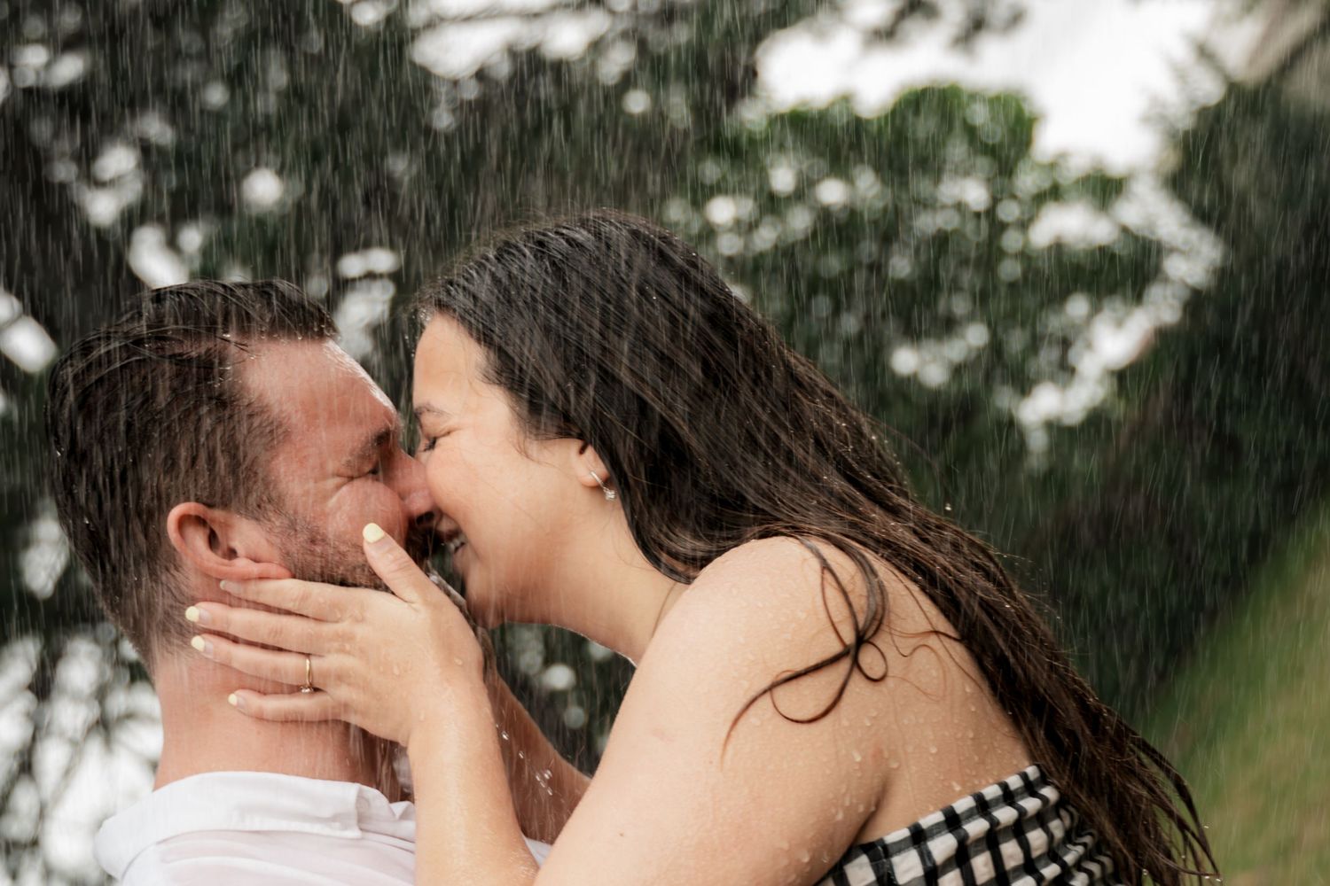Couple sharing a romantic kiss in the rain, with lush green trees in the background.