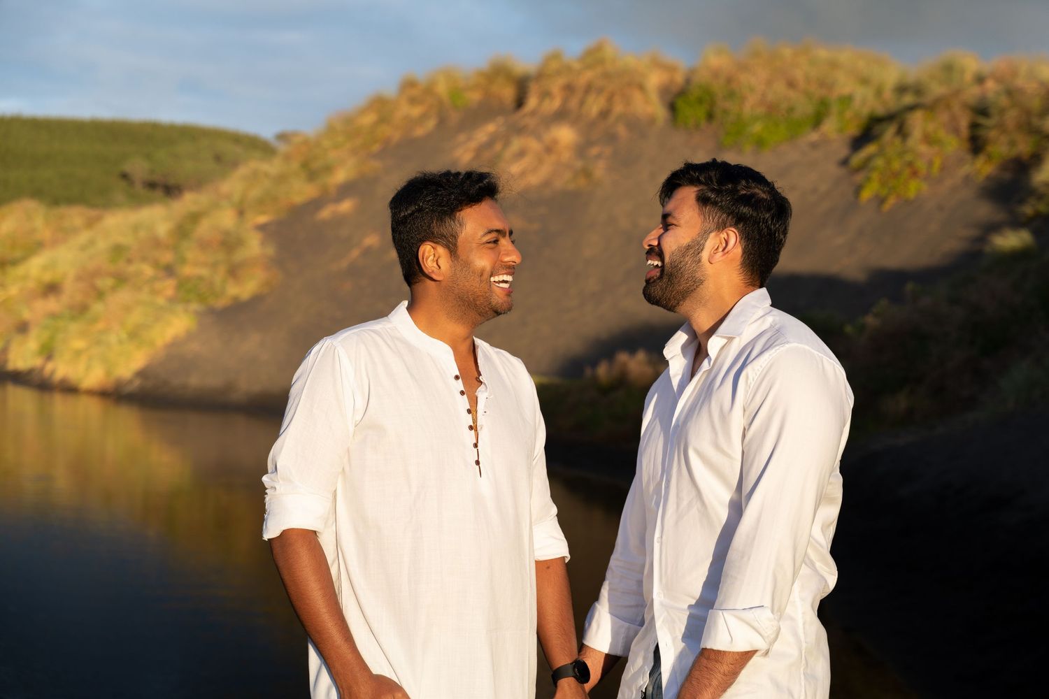 Two men in white shirts smiling at each other outdoors near a scenic lake and hillside at golden hour.