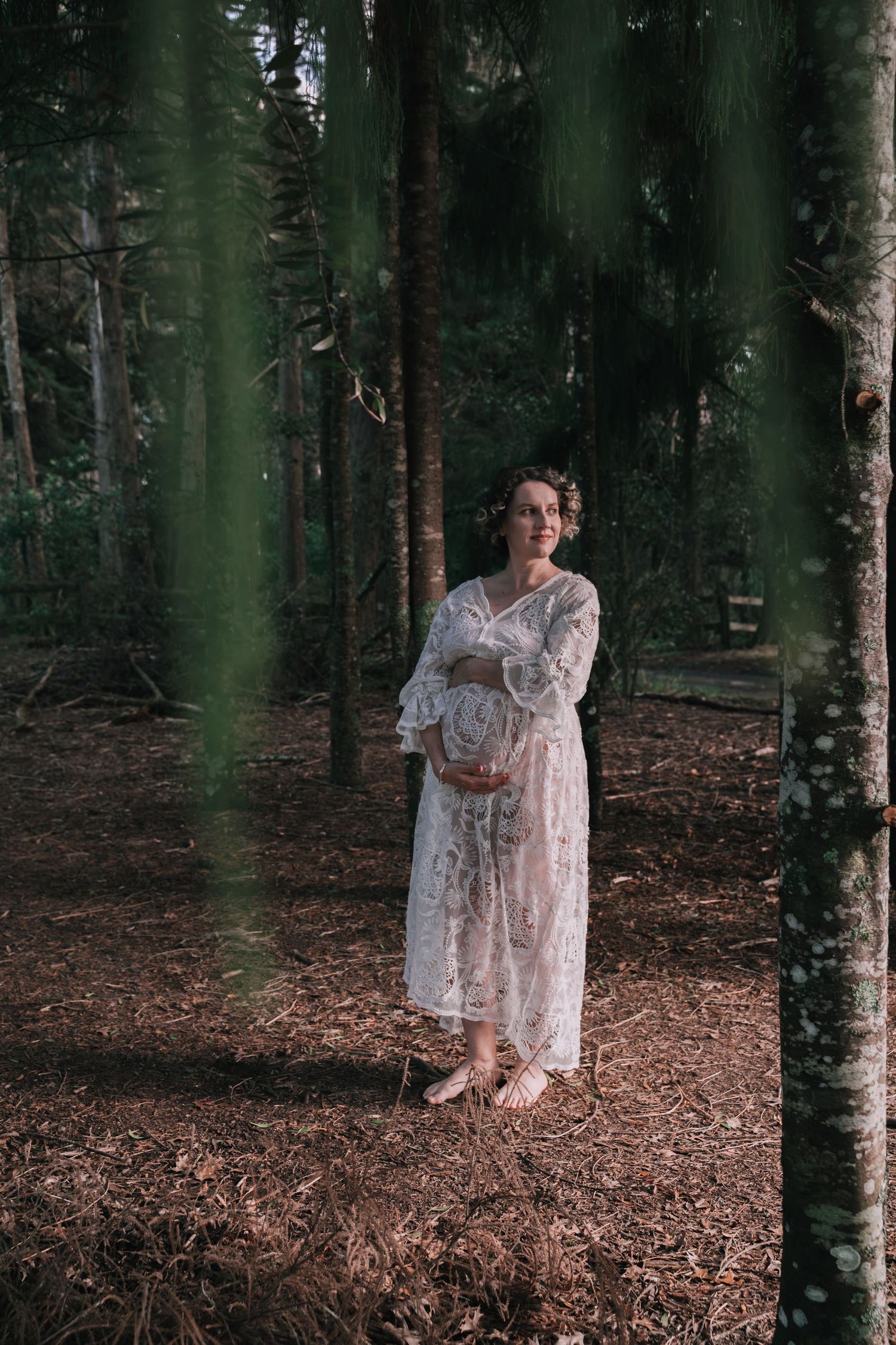 Pregnant woman in white lace dress standing barefoot in a pine forest, cradling her baby bump.