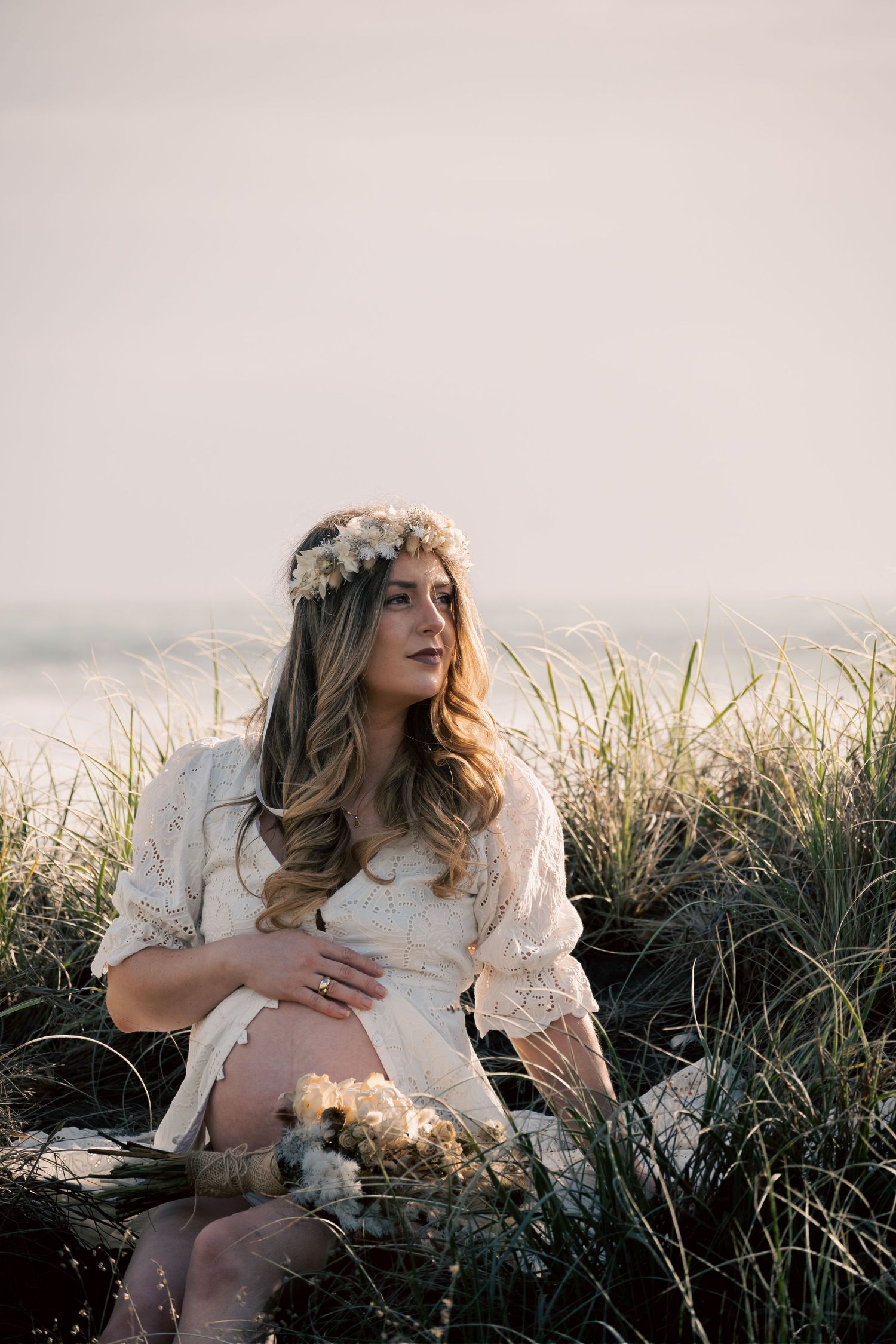 Pregnant woman in white lace dress and floral crown sits in beach grass holding bouquet at golden hour.