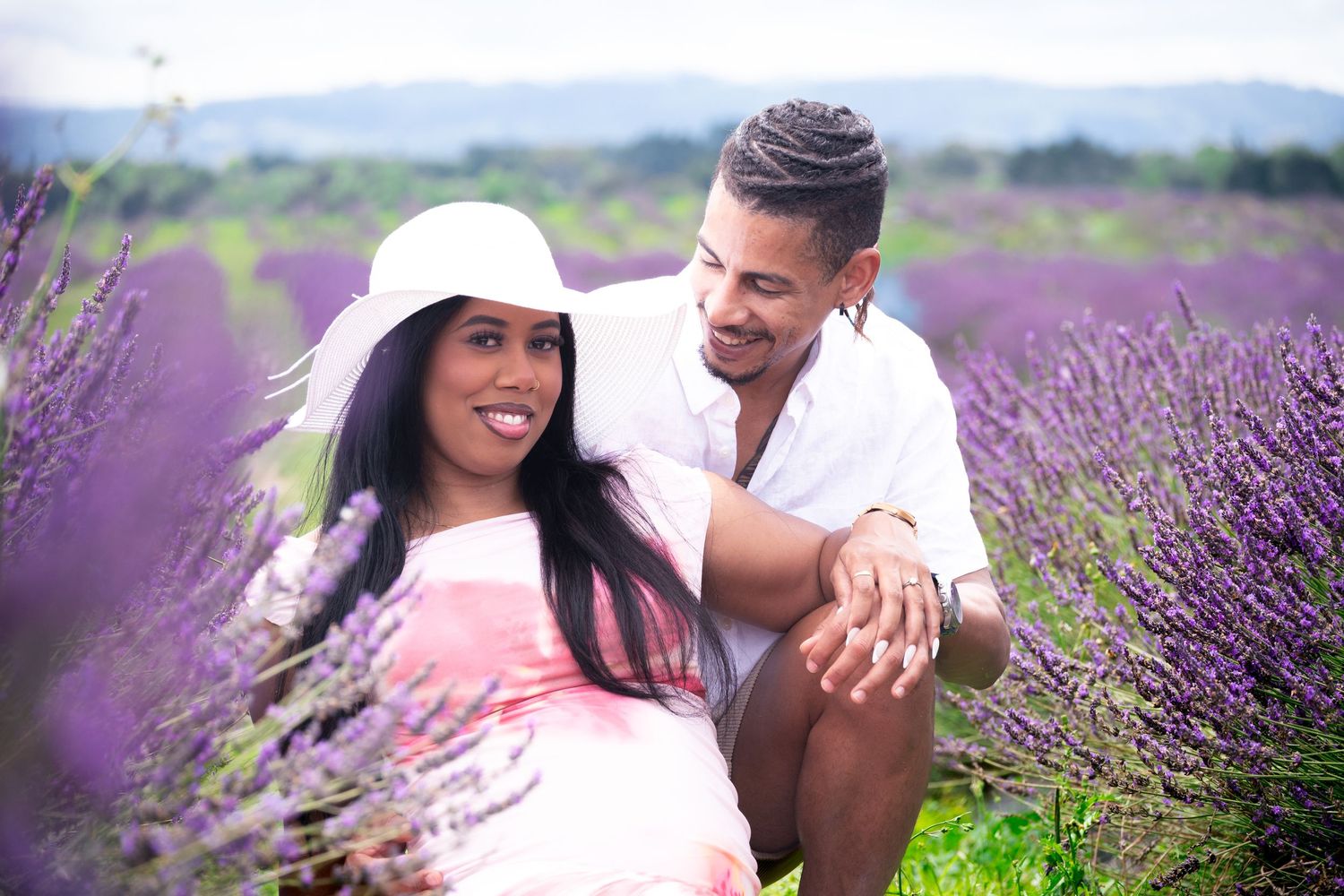 Couple sitting together in a blooming purple lavender field, woman in pink dress and white hat smiling.