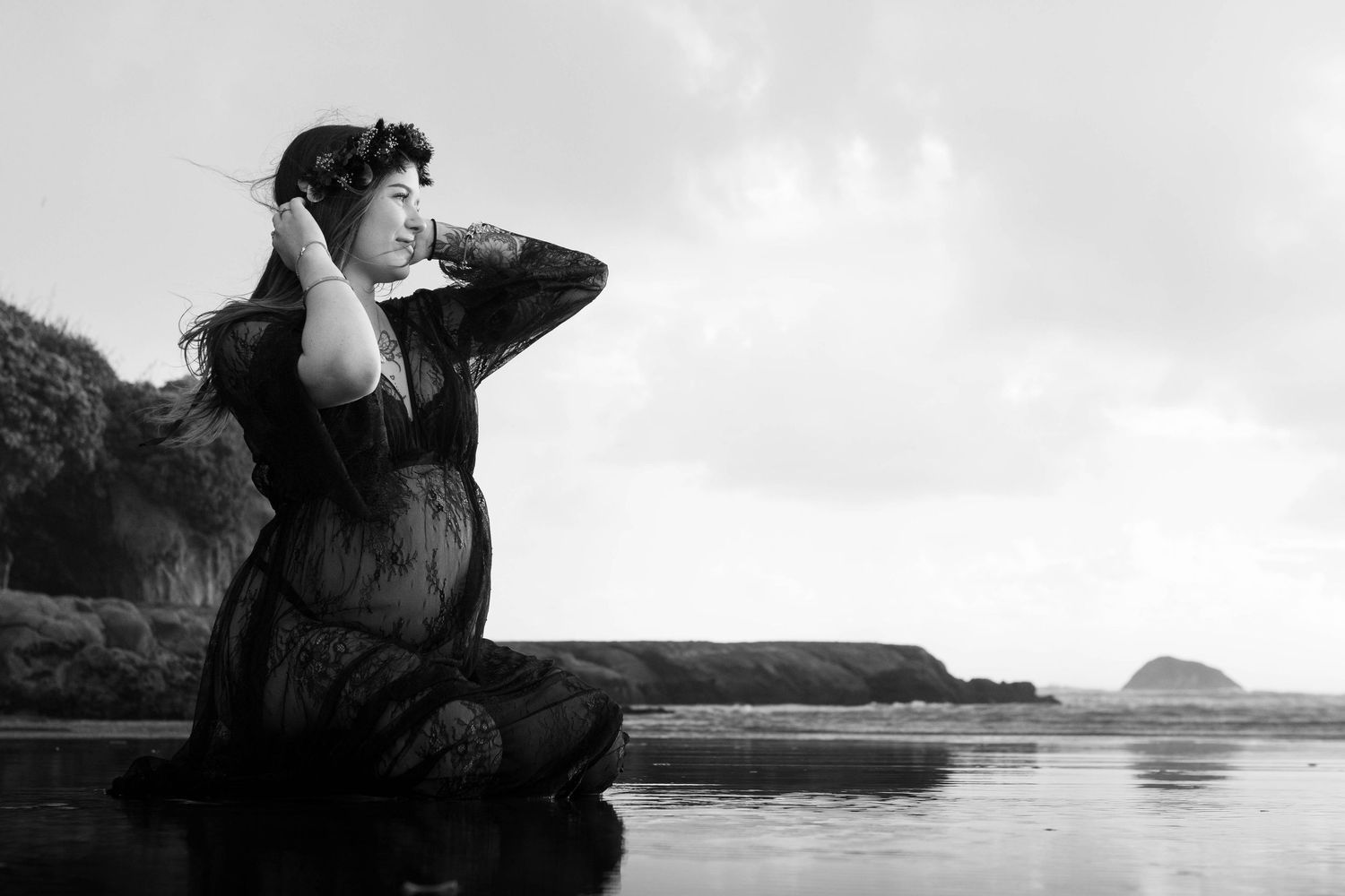 Black and white photo of a woman in a dark dress sitting by the ocean, hands in hair, with cliffs in the background.