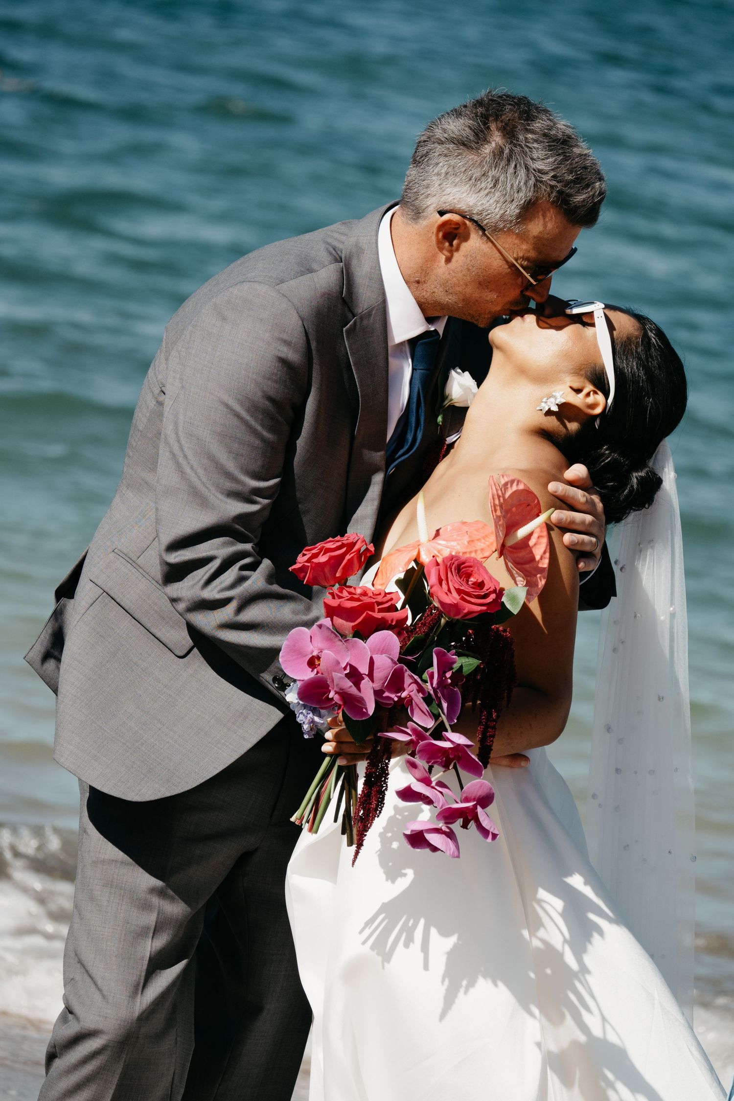 Groom in gray suit kisses bride holding colorful rose and orchid bouquet by the ocean on a north shore beach.