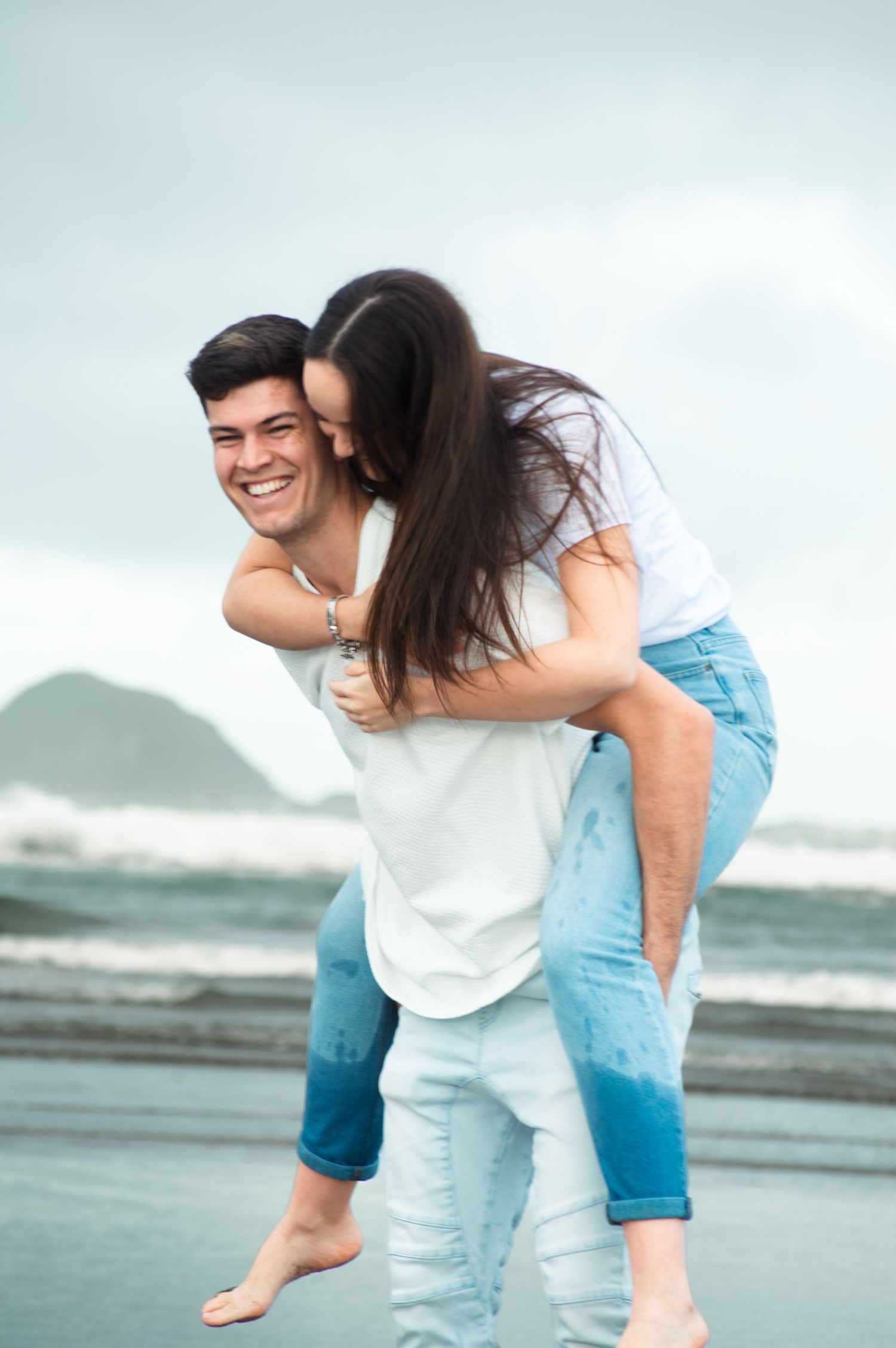 Happy couple at the beach, man giving woman a piggyback ride while she kisses his cheek, both wearing jeans.