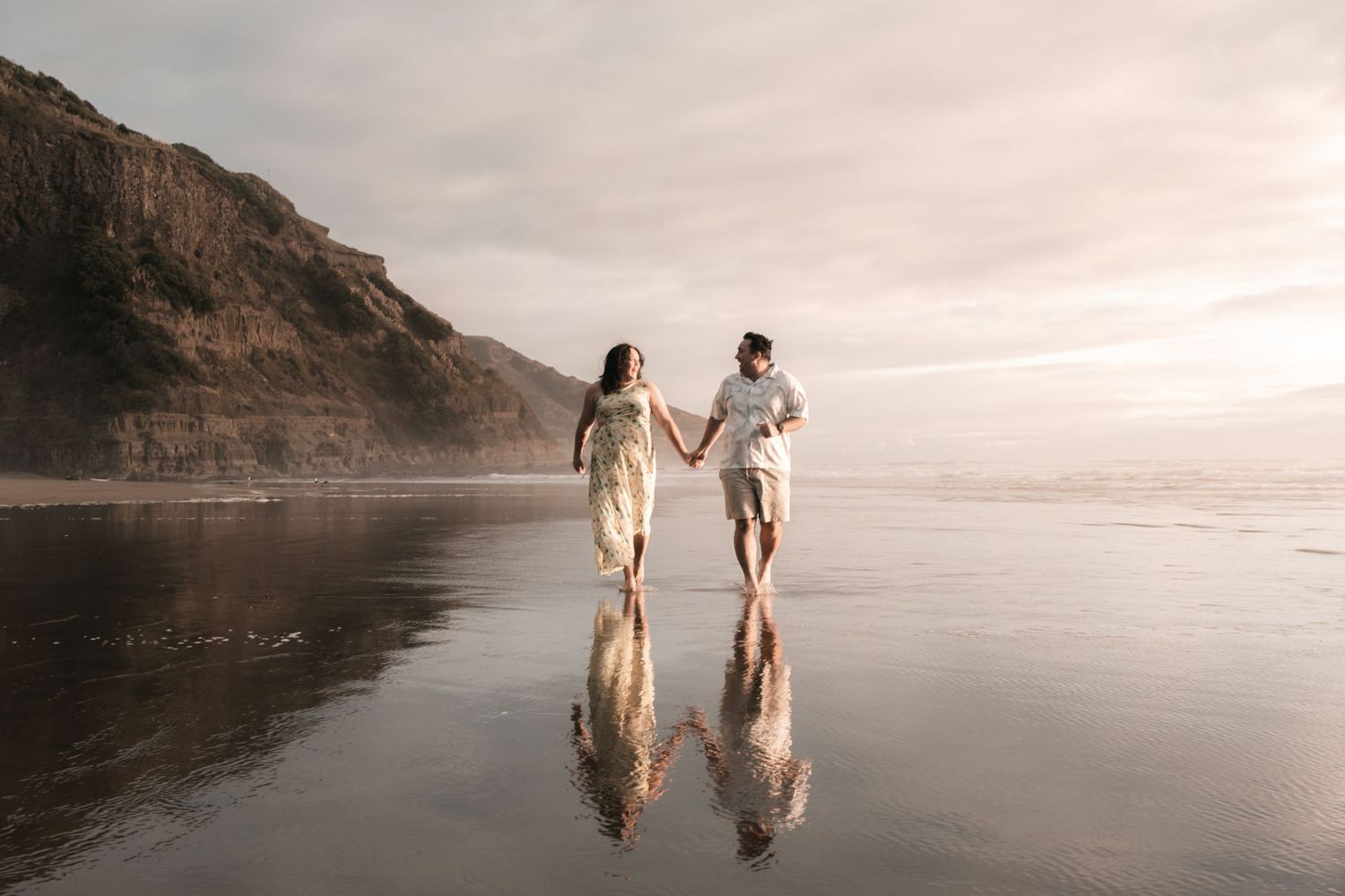 Couple holding hands walking on reflective wet beach at sunset with misty cliffs in background.