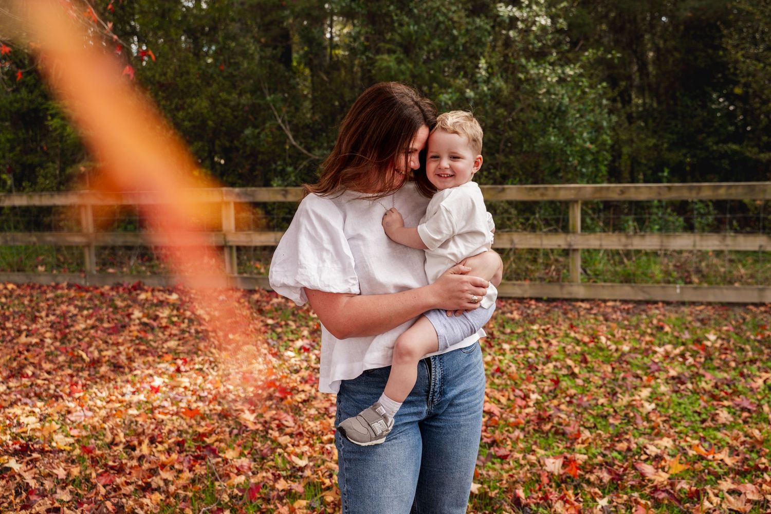 A mother holds her laughing toddler outdoors among fallen autumn leaves near a wooden fence.